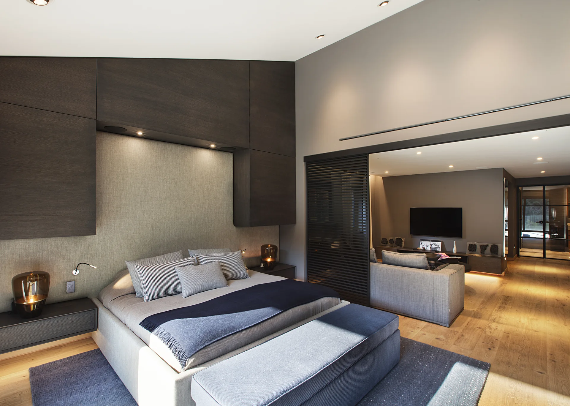 A modern bedroom features a custom dark wood headboard unit with integrated lighting next to a slatted sliding door leading to an adjoining living room.