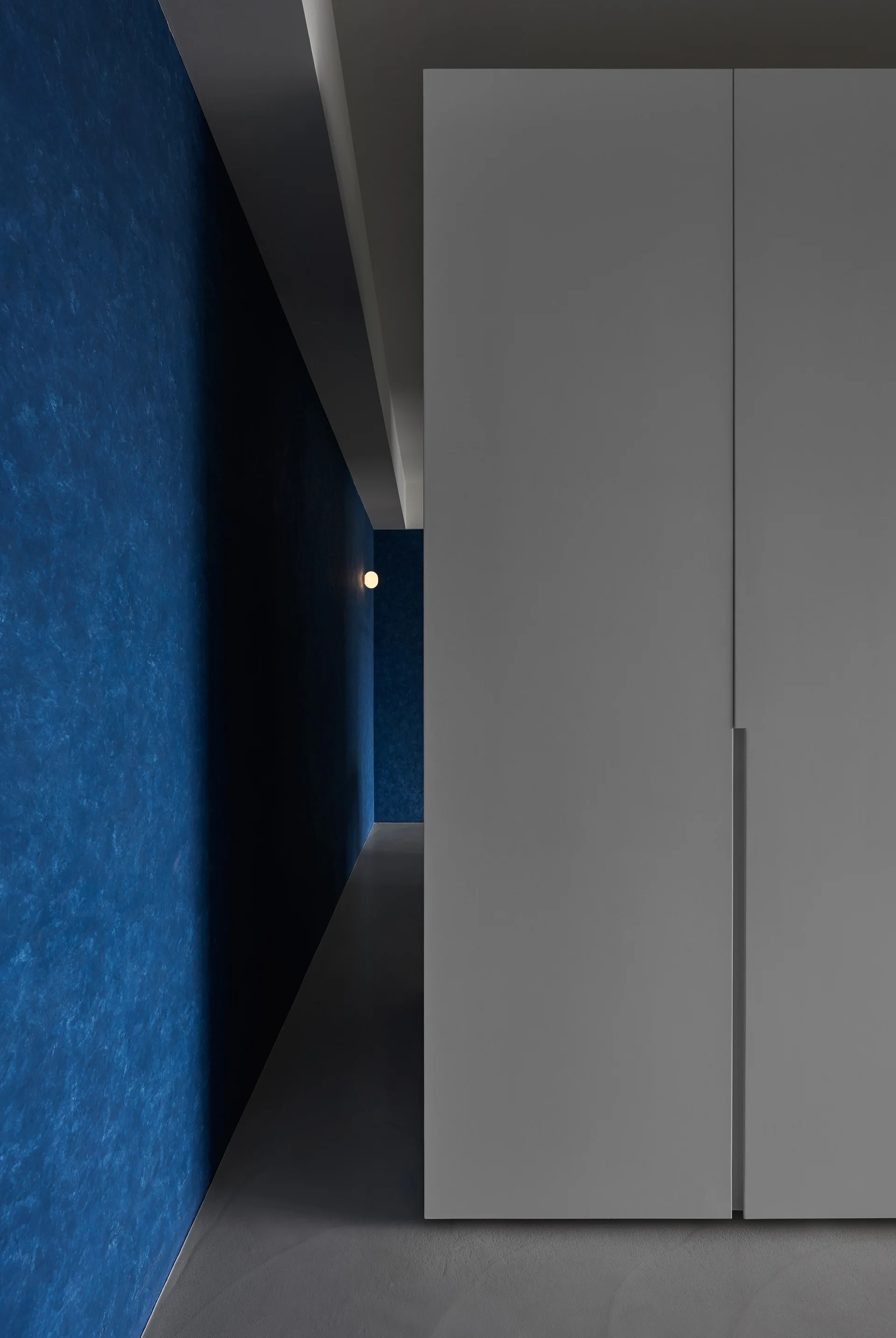 A minimalist hallway features a tall grey built-in cabinet adjacent to a deep blue textured wall illuminated by a small circular wall sconce.