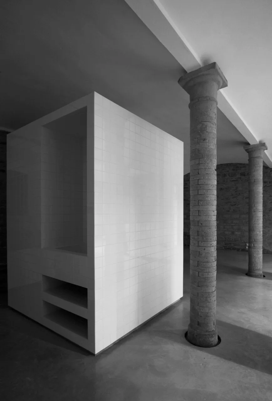 A minimalist white tiled architectural structure stands next to textured stone columns in an industrial room with concrete floors.