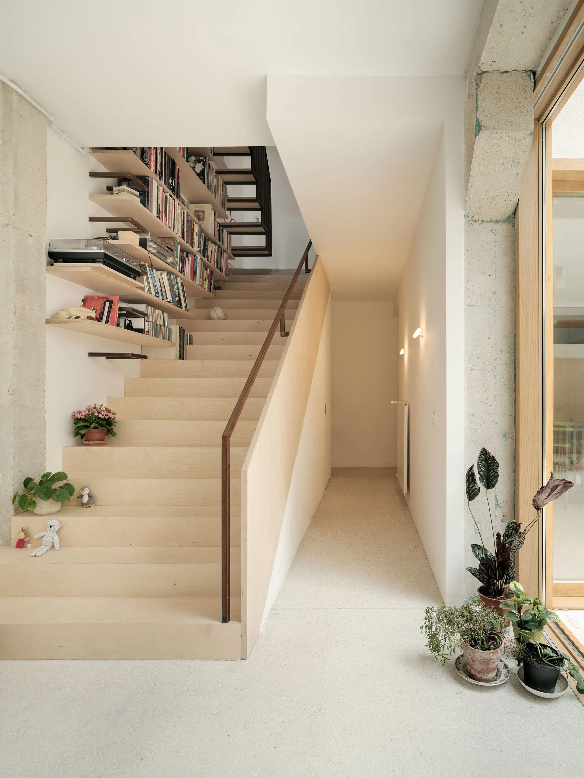 A light wood staircase featuring built-in bookshelves filled with books on the left, next to an exposed concrete column and a hallway.