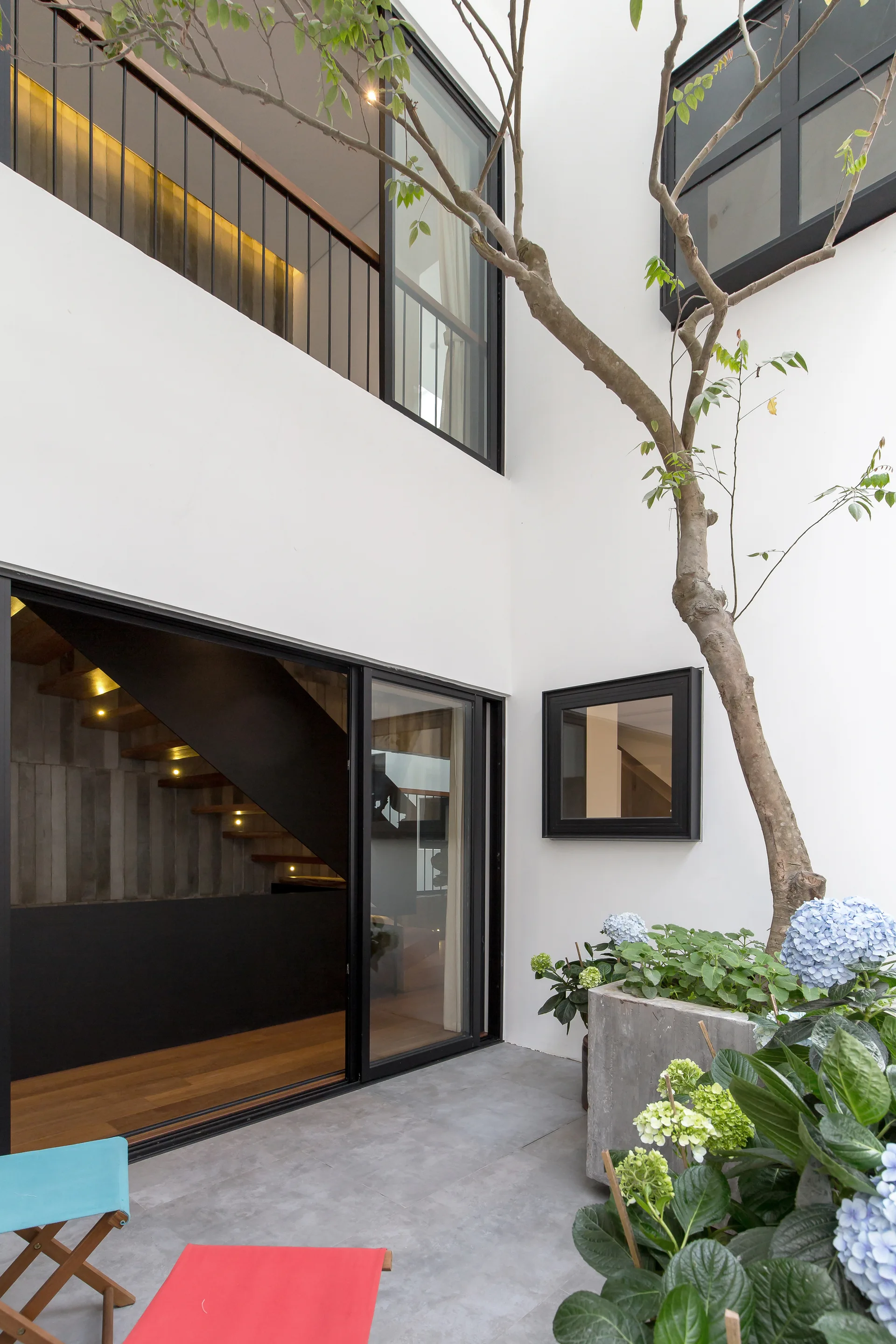 A modern white courtyard features large black-framed sliding glass doors revealing an interior staircase, concrete flooring, and a stone planter.