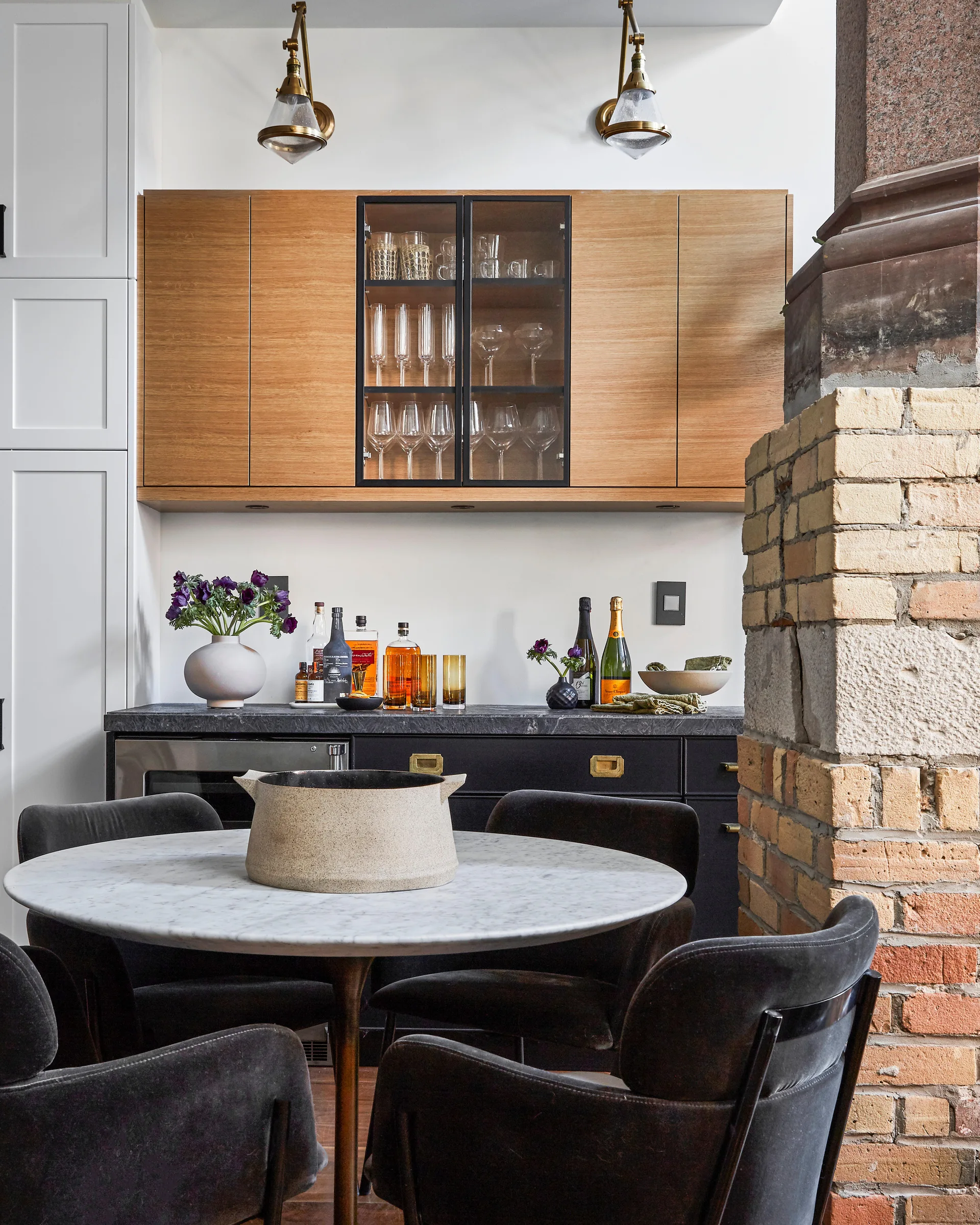 A modern home bar featuring wood upper cabinets, dark base cabinetry, and a round marble dining table with dark upholstered chairs.