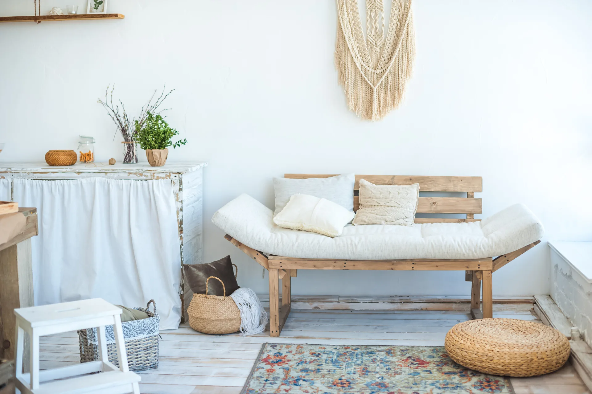 A rustic wooden bench with cream cushions sits against a white wall below a macrame hanging, next to a white skirted console.