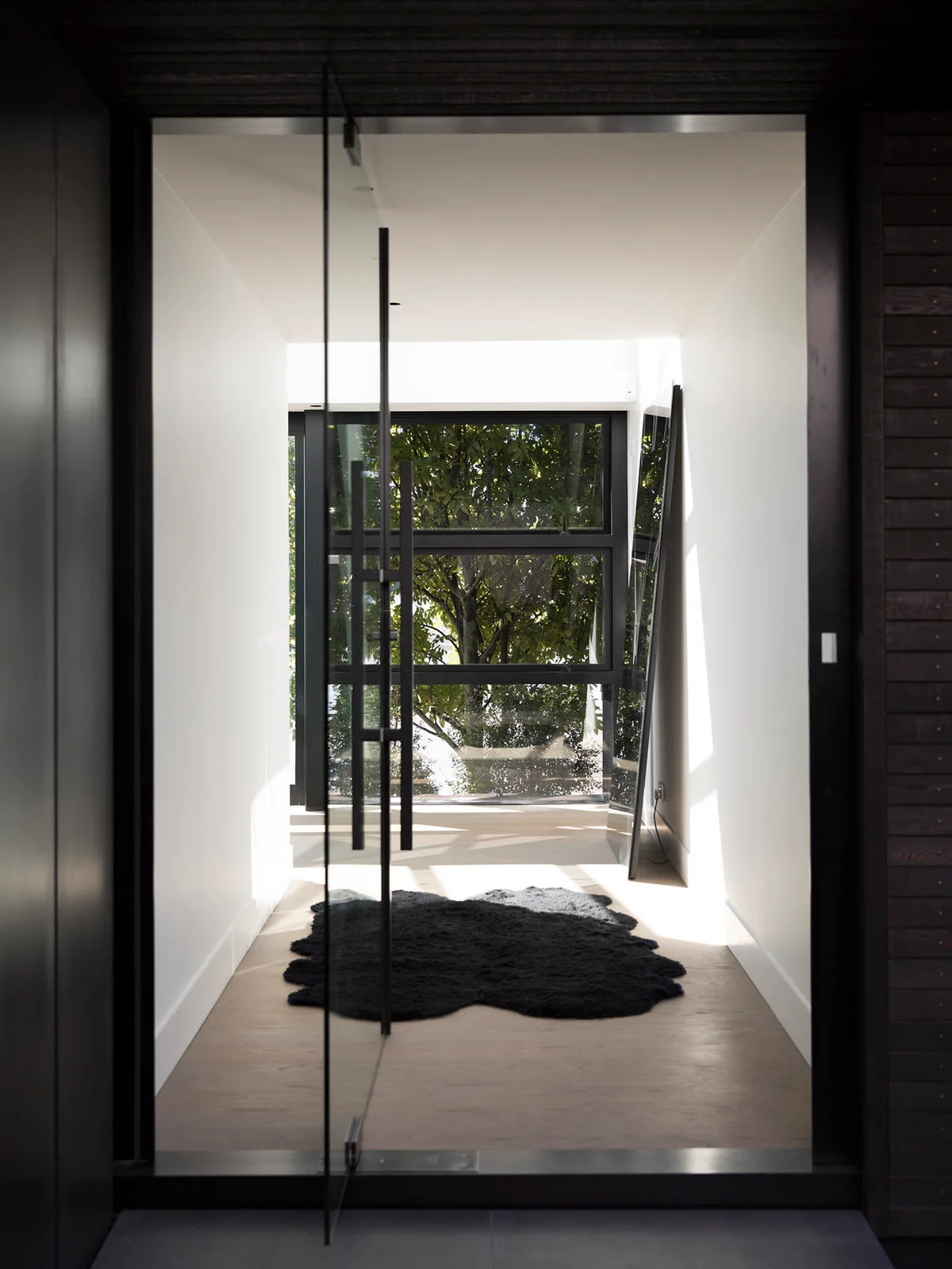 A bright modern entryway seen through a glass door, featuring white walls, light wood floors, a black hide rug, and a tall leaning mirror.