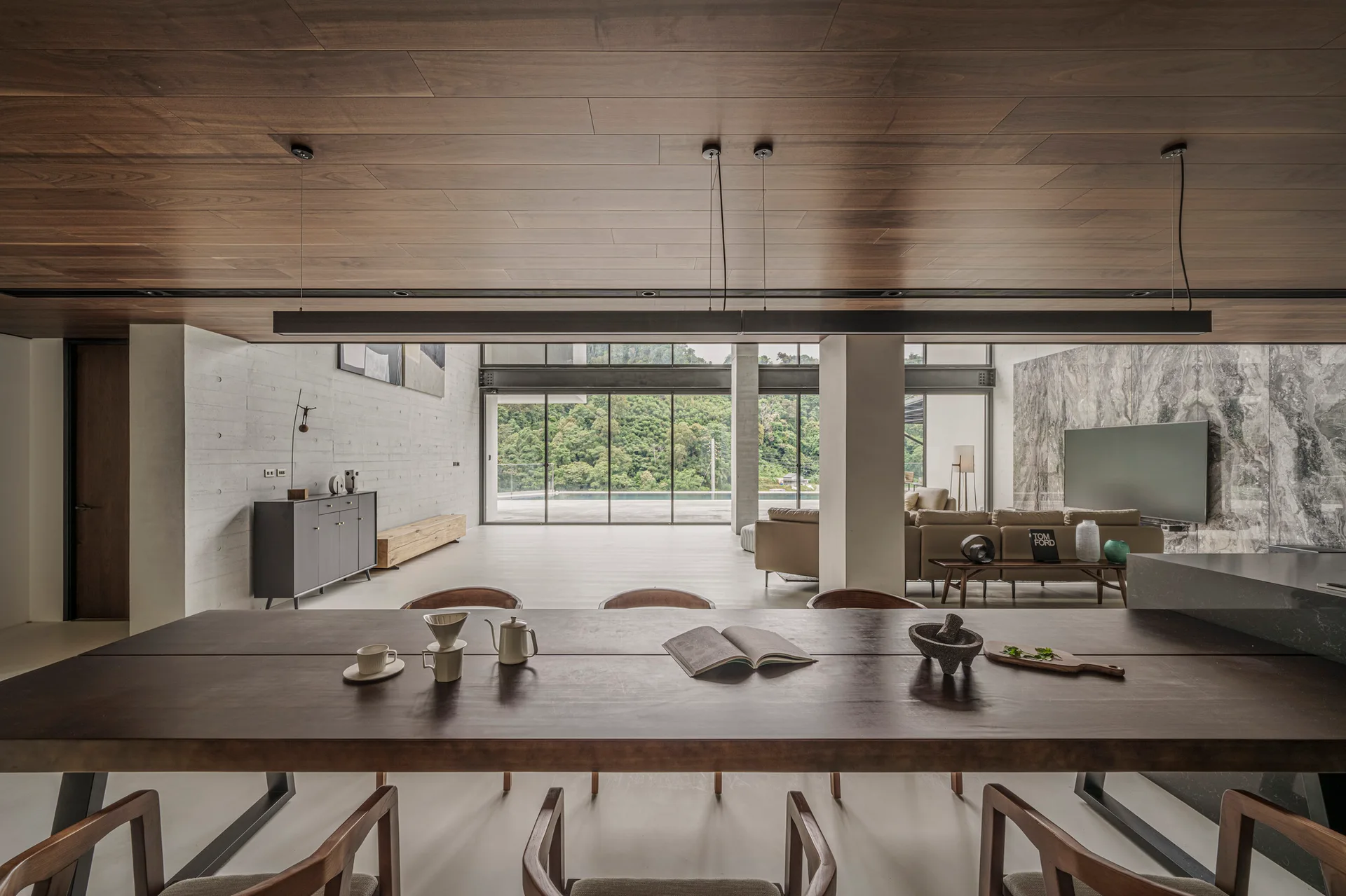 A long wood dining table stands beneath a paneled wood ceiling in a modern open-concept space featuring concrete walls and large windows.