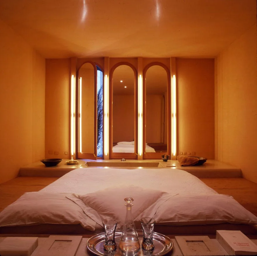A low platform bed with white sheets sits in a minimalist warm orange bedroom before a wall featuring three arched mirrors with vertical tube lights.