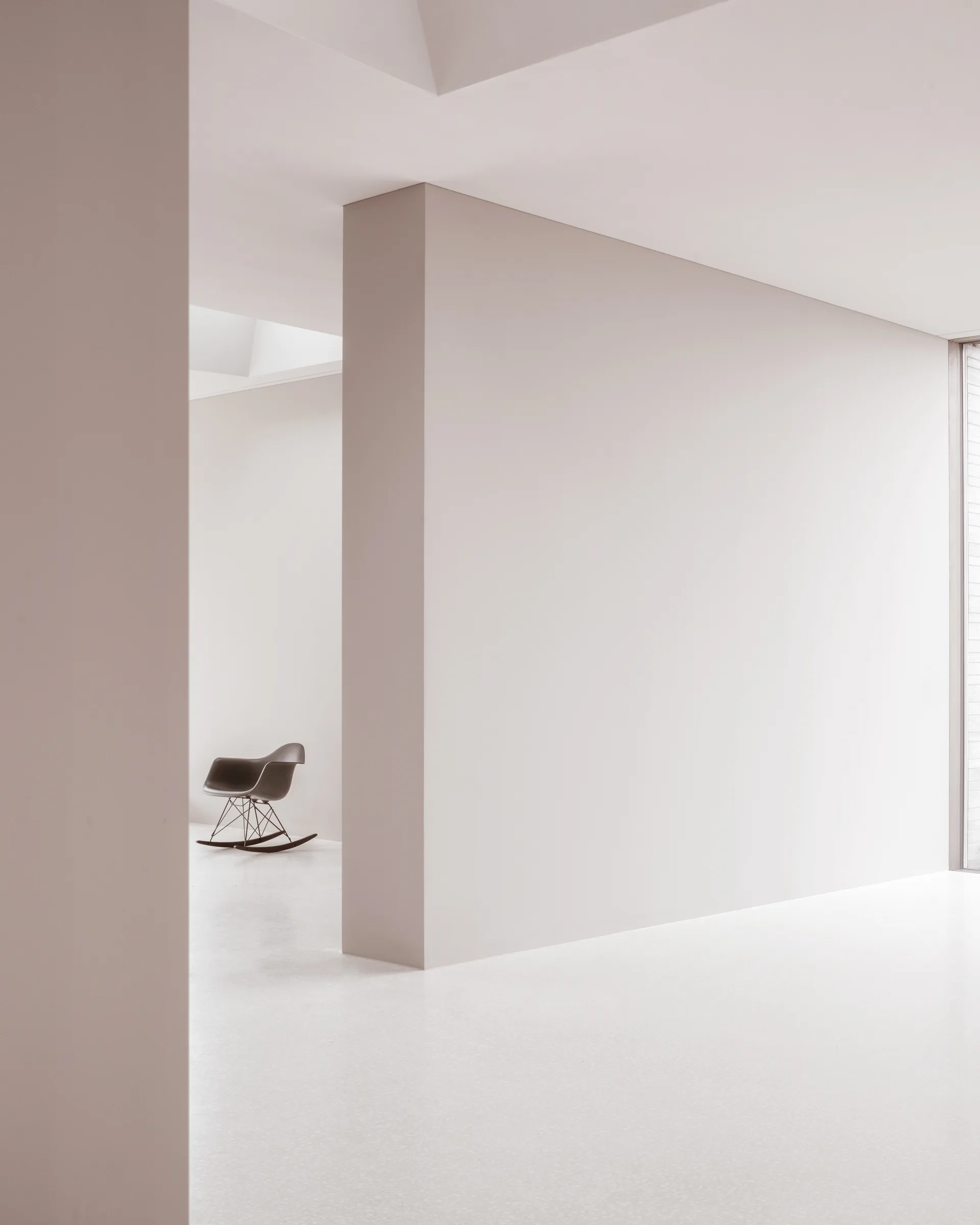 A minimalist white room features clean geometric partition walls and a single black rocking chair visible in the background.