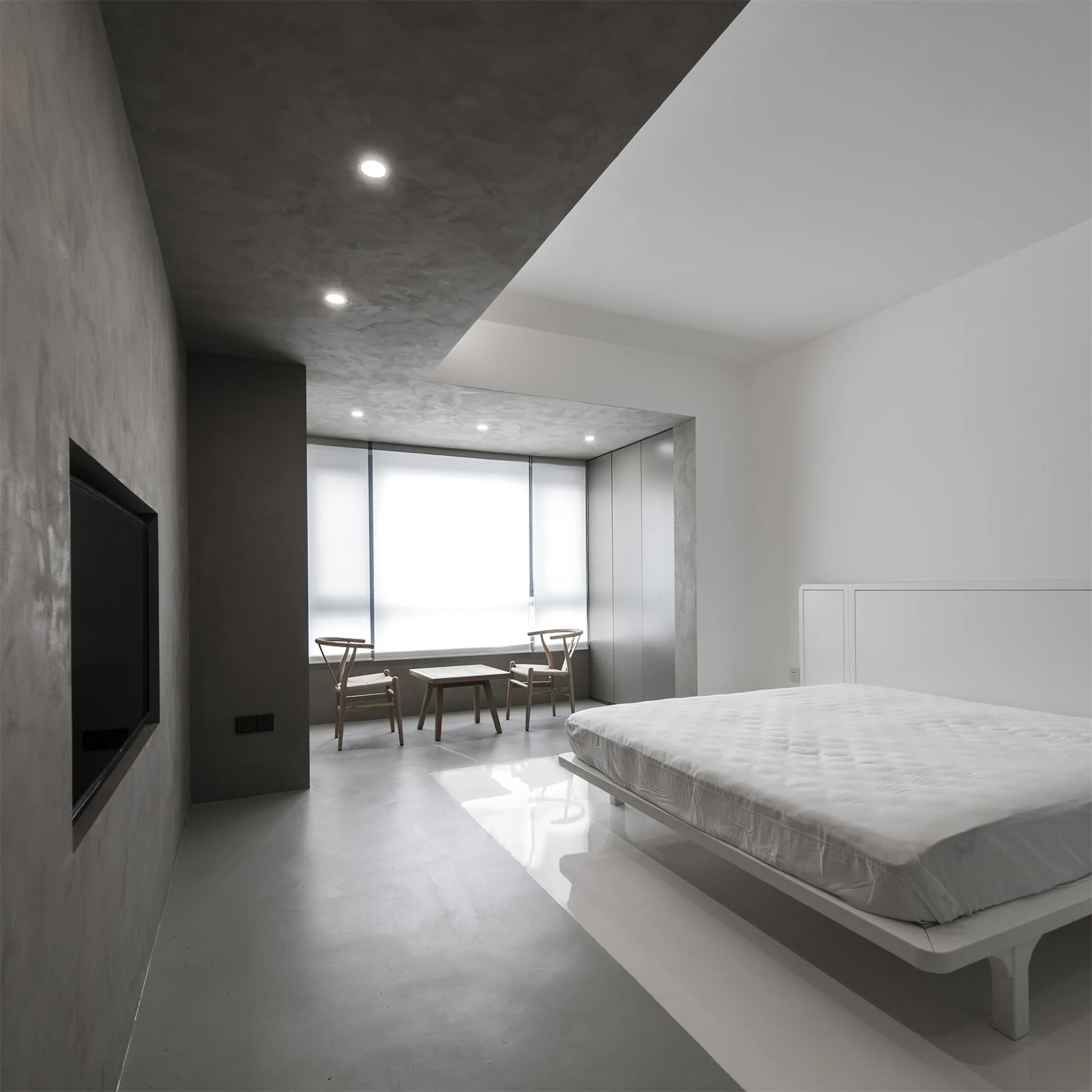 A minimalist bedroom features a white platform bed, a seating area near a large window, and a contrasting grey textured plaster wall and ceiling.