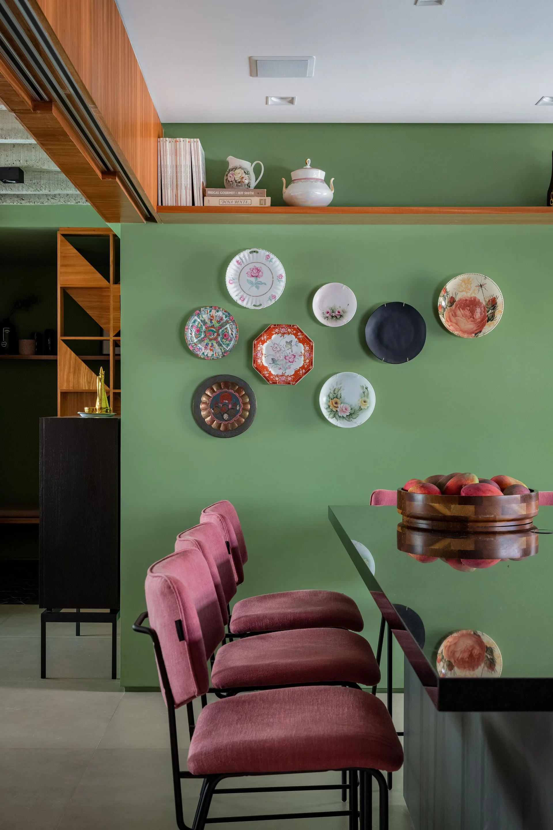 A green dining room wall features a gallery of patterned decorative plates above dusty pink chairs and a glossy black table.