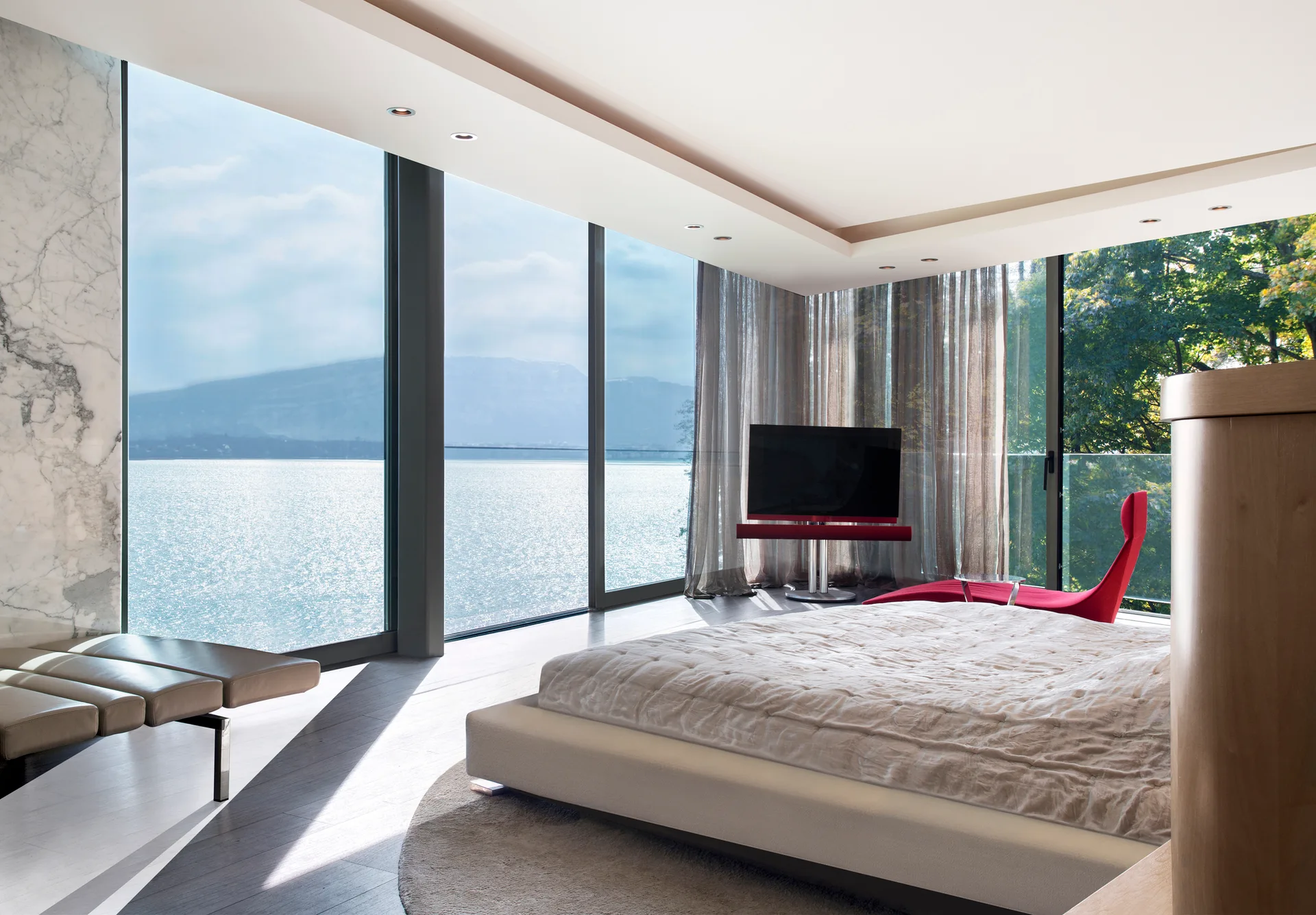 A minimalist modern bedroom features a low-profile white bed, a red lounge chair, and floor-to-ceiling windows overlooking a lake.
