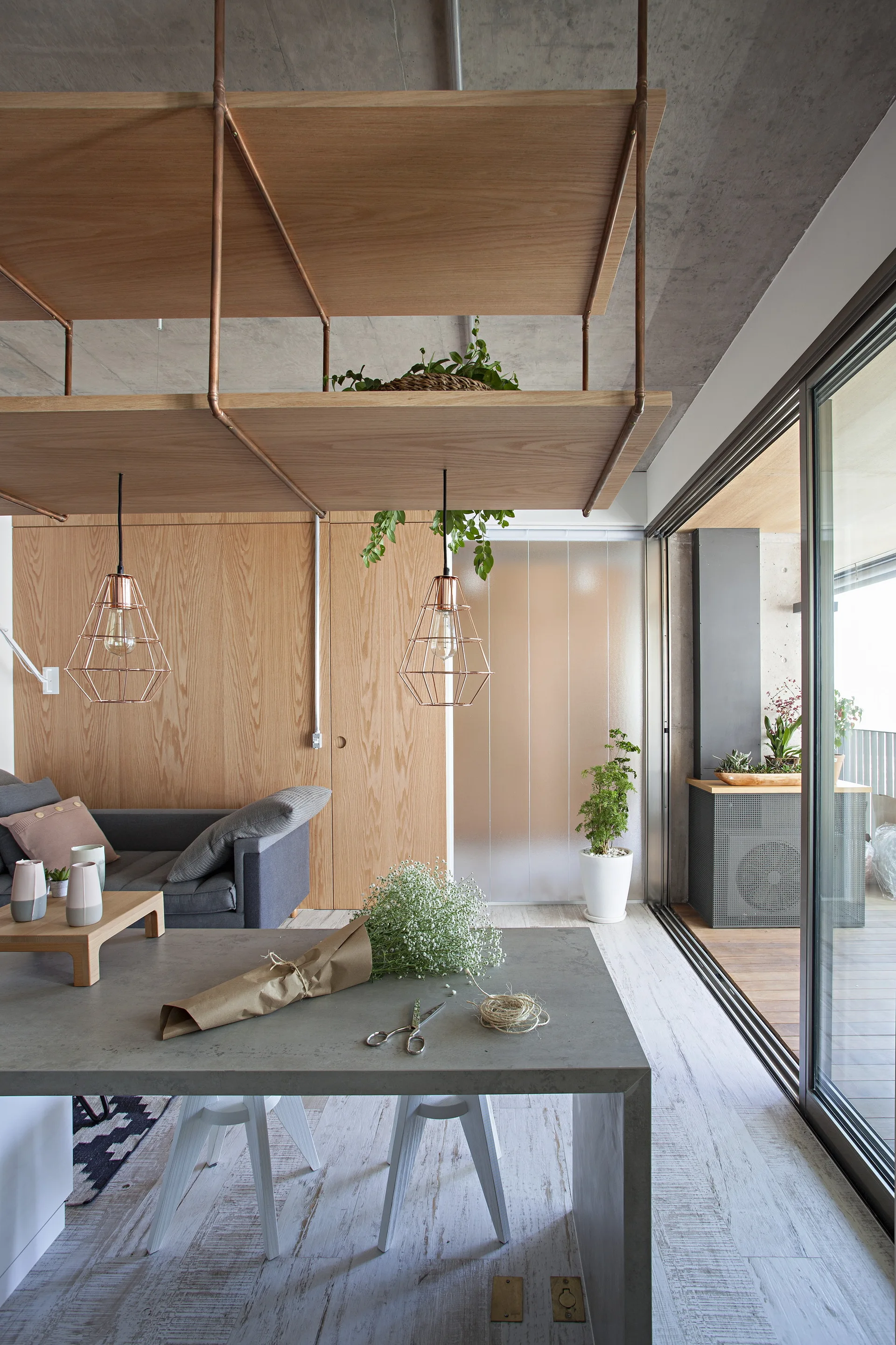 A contemporary living room features a suspended wooden shelf hanging from a concrete ceiling, wood-paneled walls, a gray sofa, and sliding glass doors.