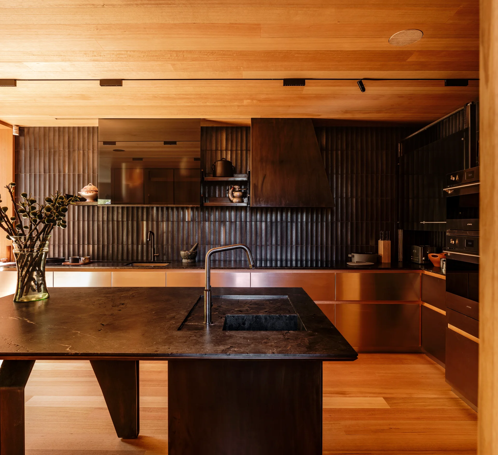 A modern kitchen features a dark stone island, brushed copper cabinets, a vertical ribbed tile backsplash, and a warm wood plank ceiling.