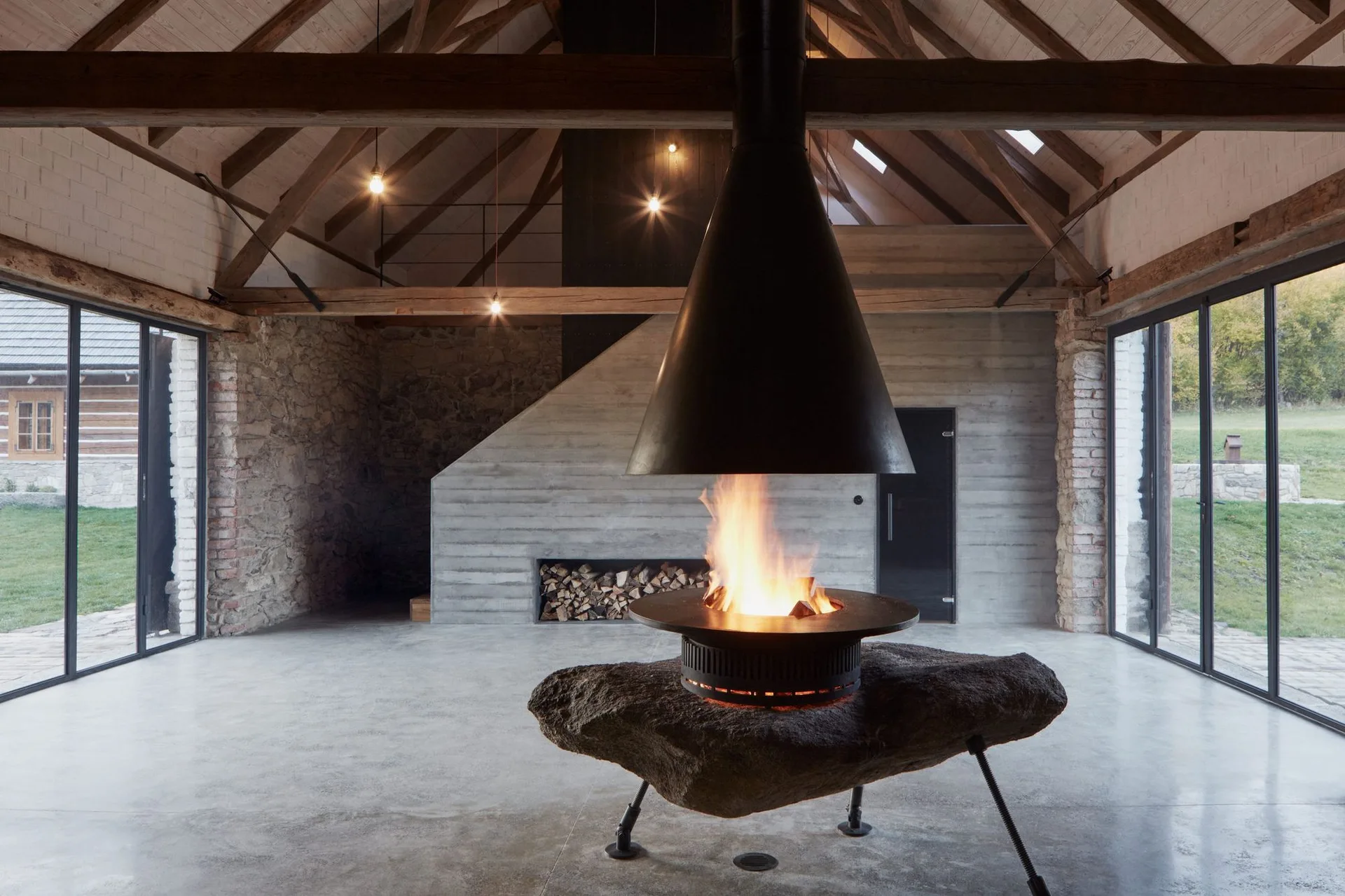 A rustic-modern room features a vaulted wood ceiling, concrete walls, and a central suspended metal fireplace supported by a large natural stone boulder.