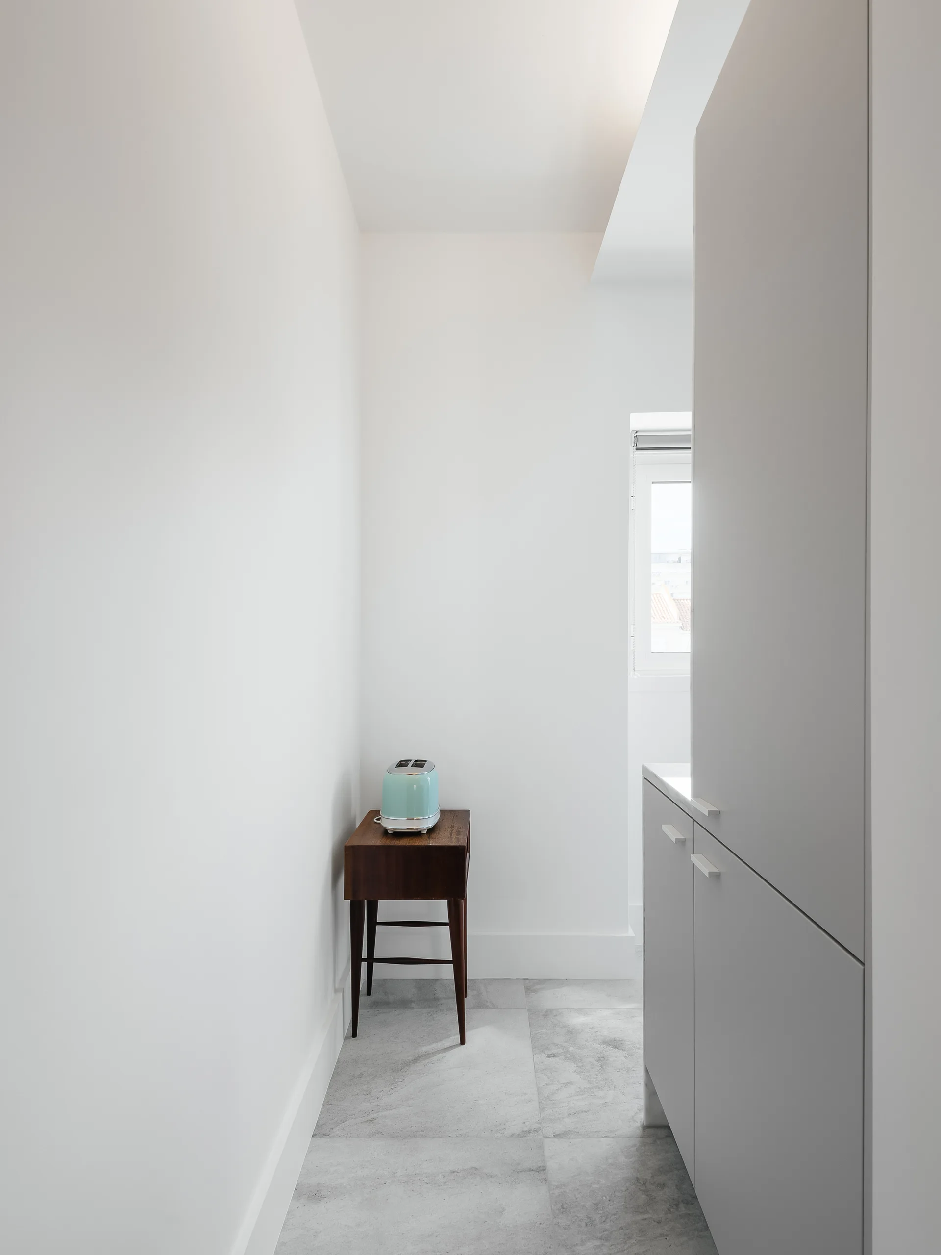 A minimalist kitchen corner features white built-in cabinetry, grey stone-like floor tiles, and a small dark wood console table with a blue toaster.