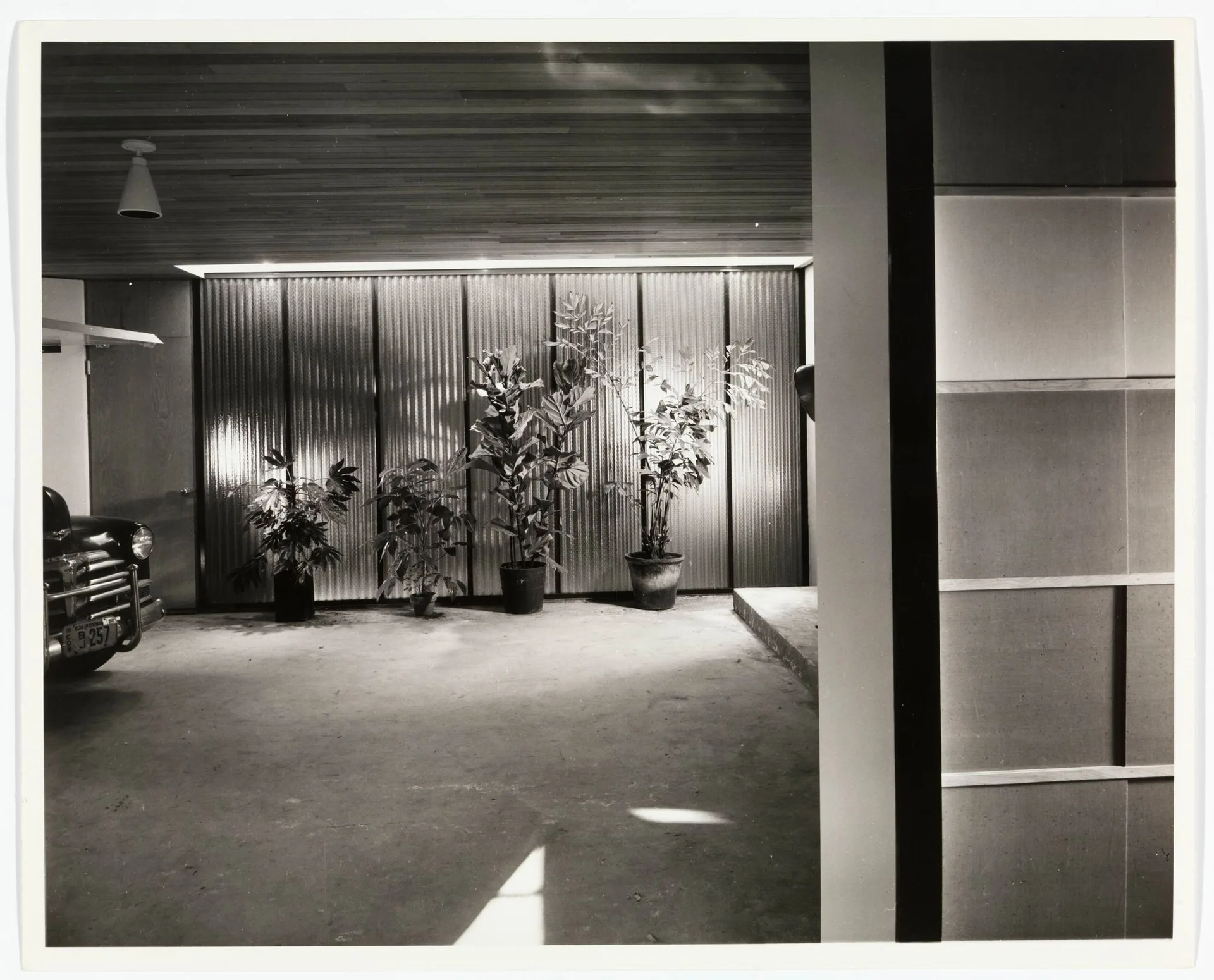 A vintage car parked in a mid-century modern garage featuring a ribbed metal wall, slatted wood ceiling, and several potted leafy plants.