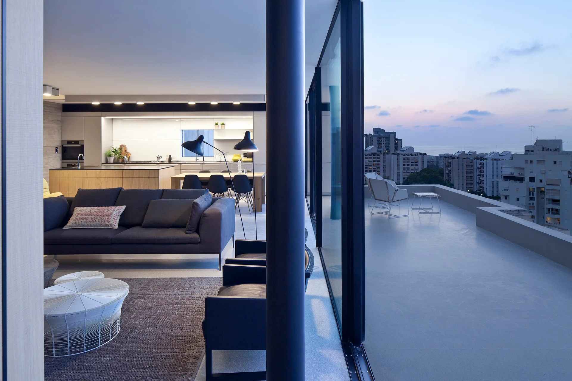 A modern open-plan living room with a dark grey sofa and glass doors opening onto a large balcony overlooking a city skyline at dusk.