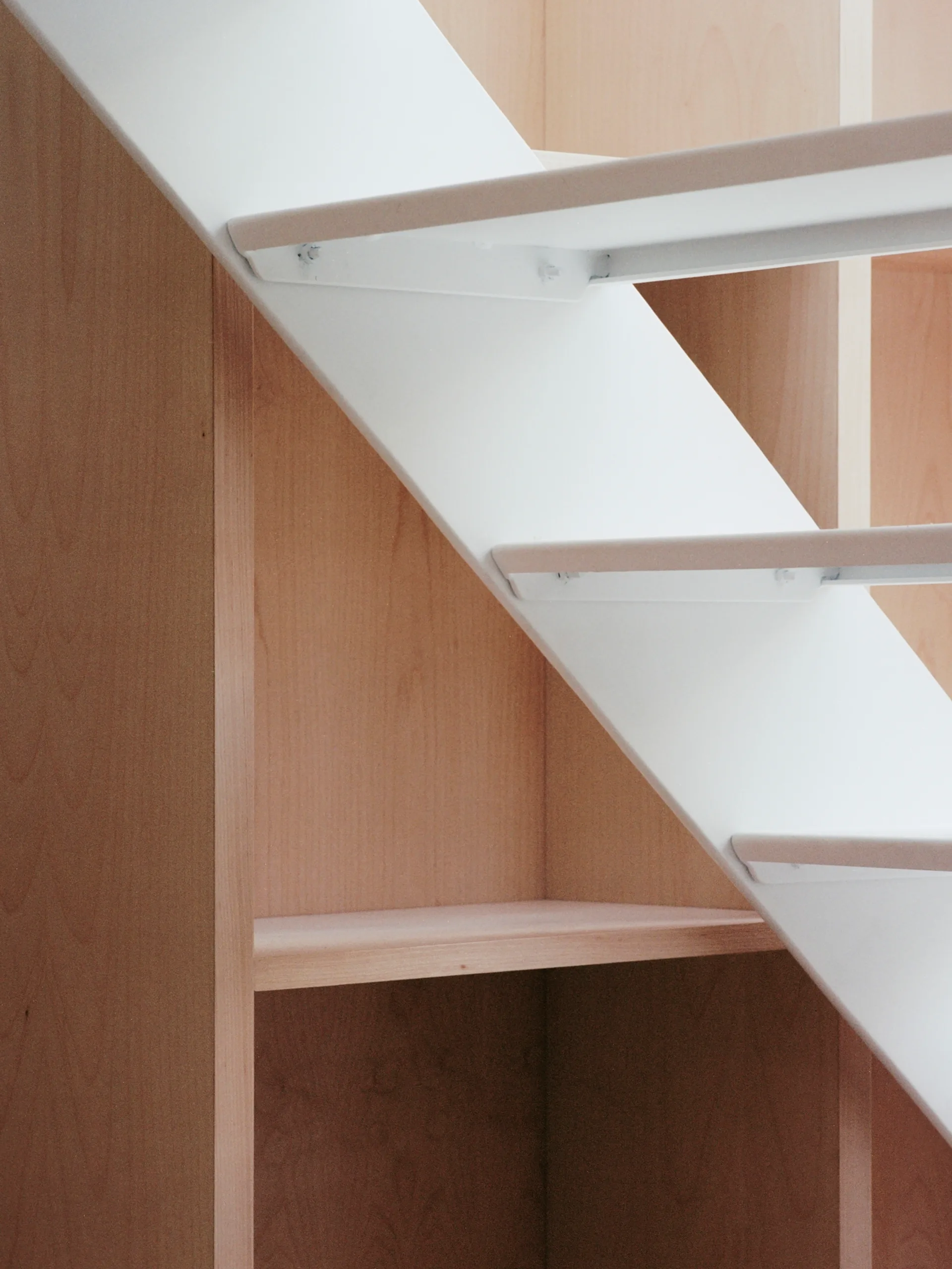 A close-up shot of a white metal staircase structure featuring integrated light wood shelving and cabinetry built directly underneath the stairs.