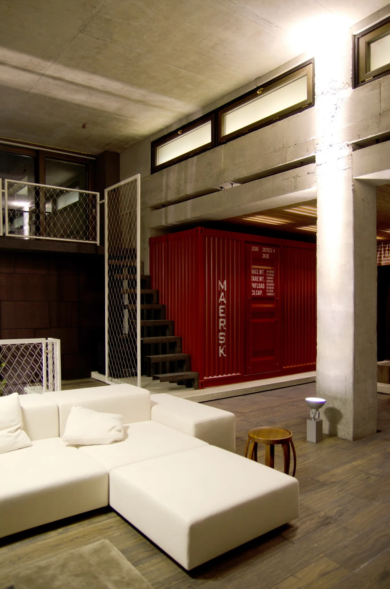 An industrial loft living room with a white modular sectional sofa, a large red shipping container, raw concrete walls, and dark wood floors.