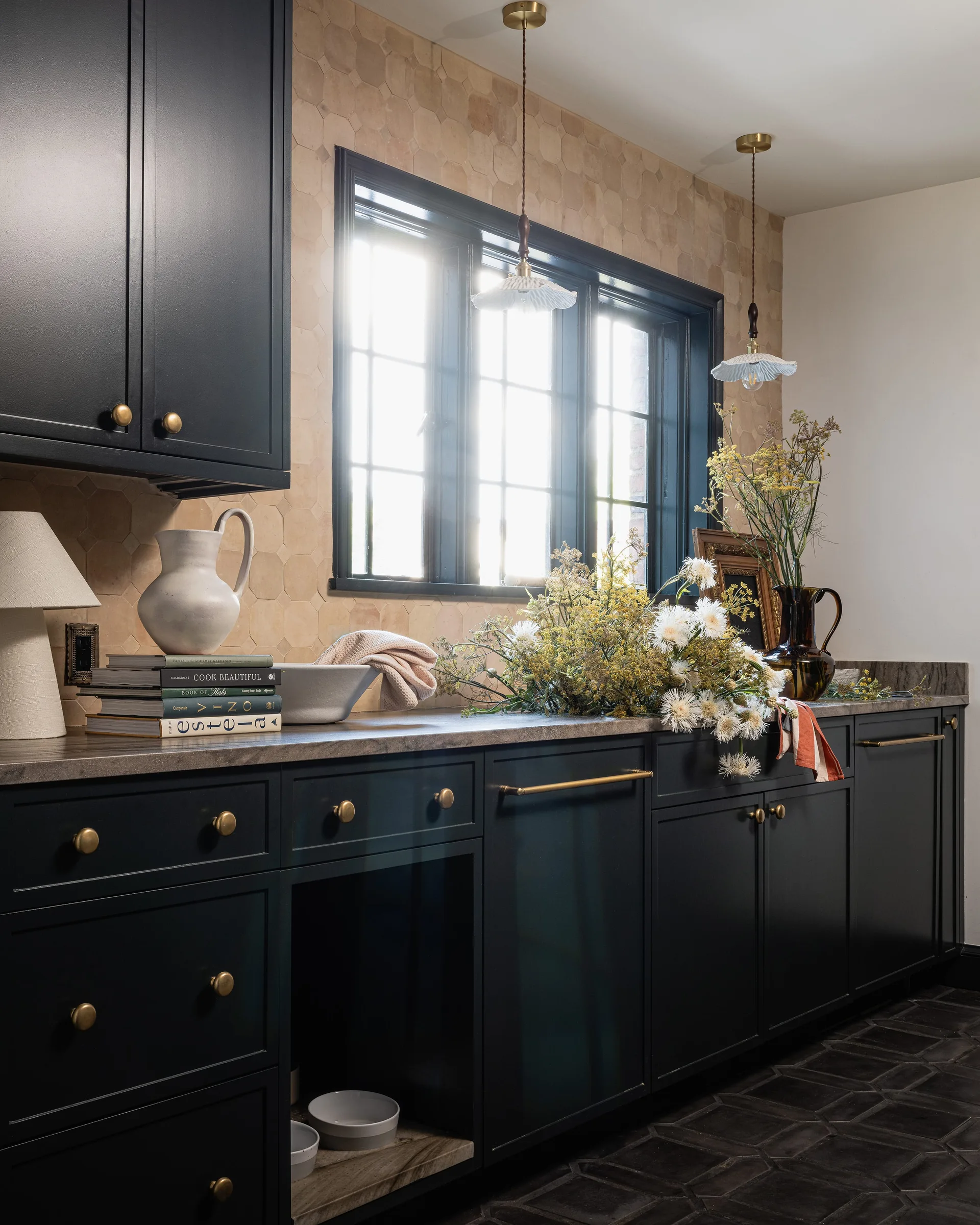 Dark charcoal kitchen cabinets are paired with a hexagonal terracotta tile backsplash, a stone countertop, and two hanging scalloped pendant lights.