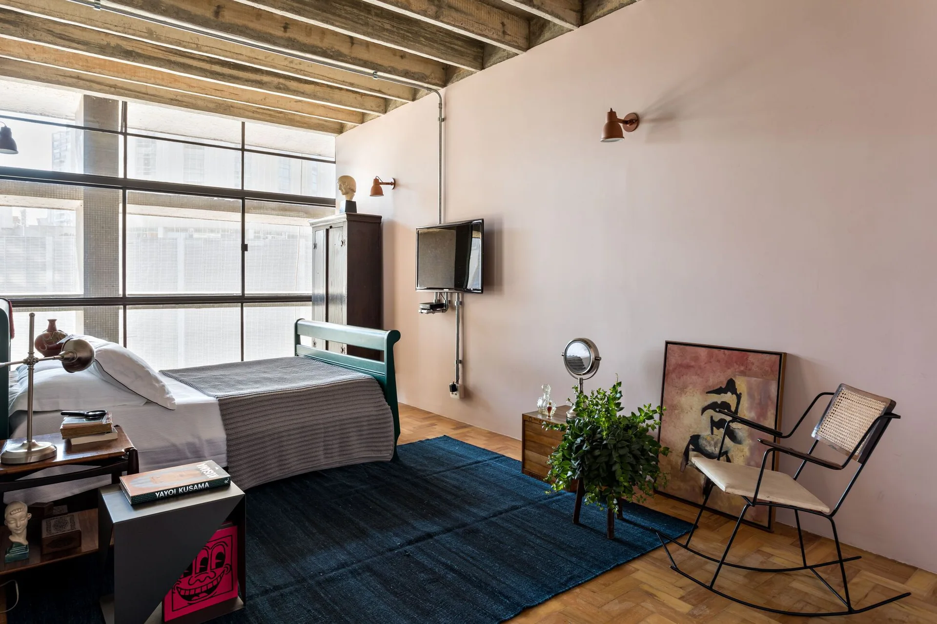 A bedroom features light pink walls, exposed concrete ceiling beams, parquet flooring, a green bed frame, and a dark blue rug.