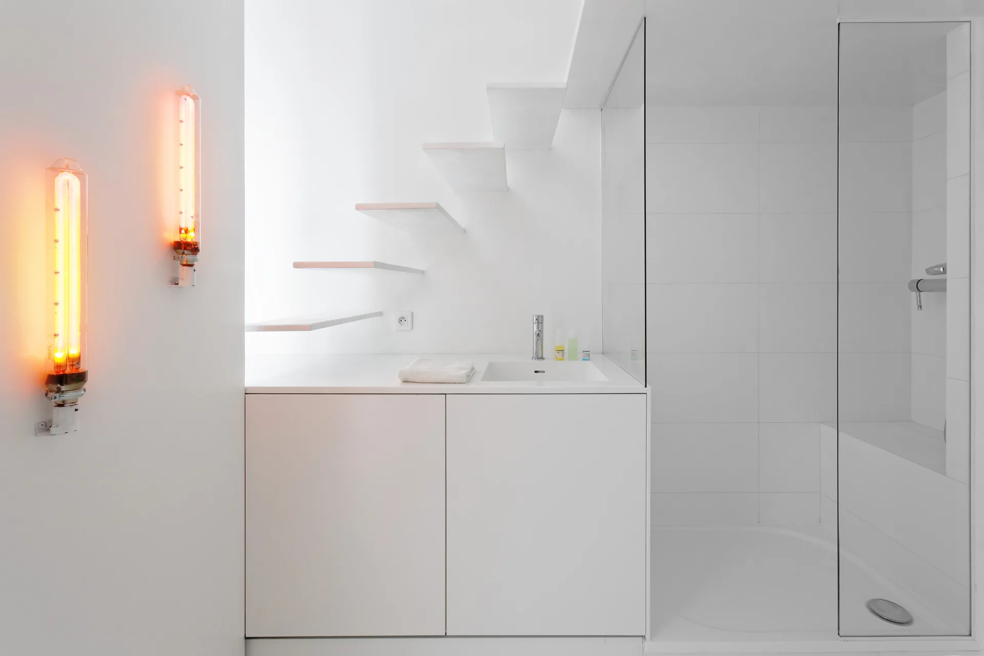 A minimalist white bathroom features a floating vanity, a glass shower stall, floating stair treads, and two vertical orange glowing wall sconces.