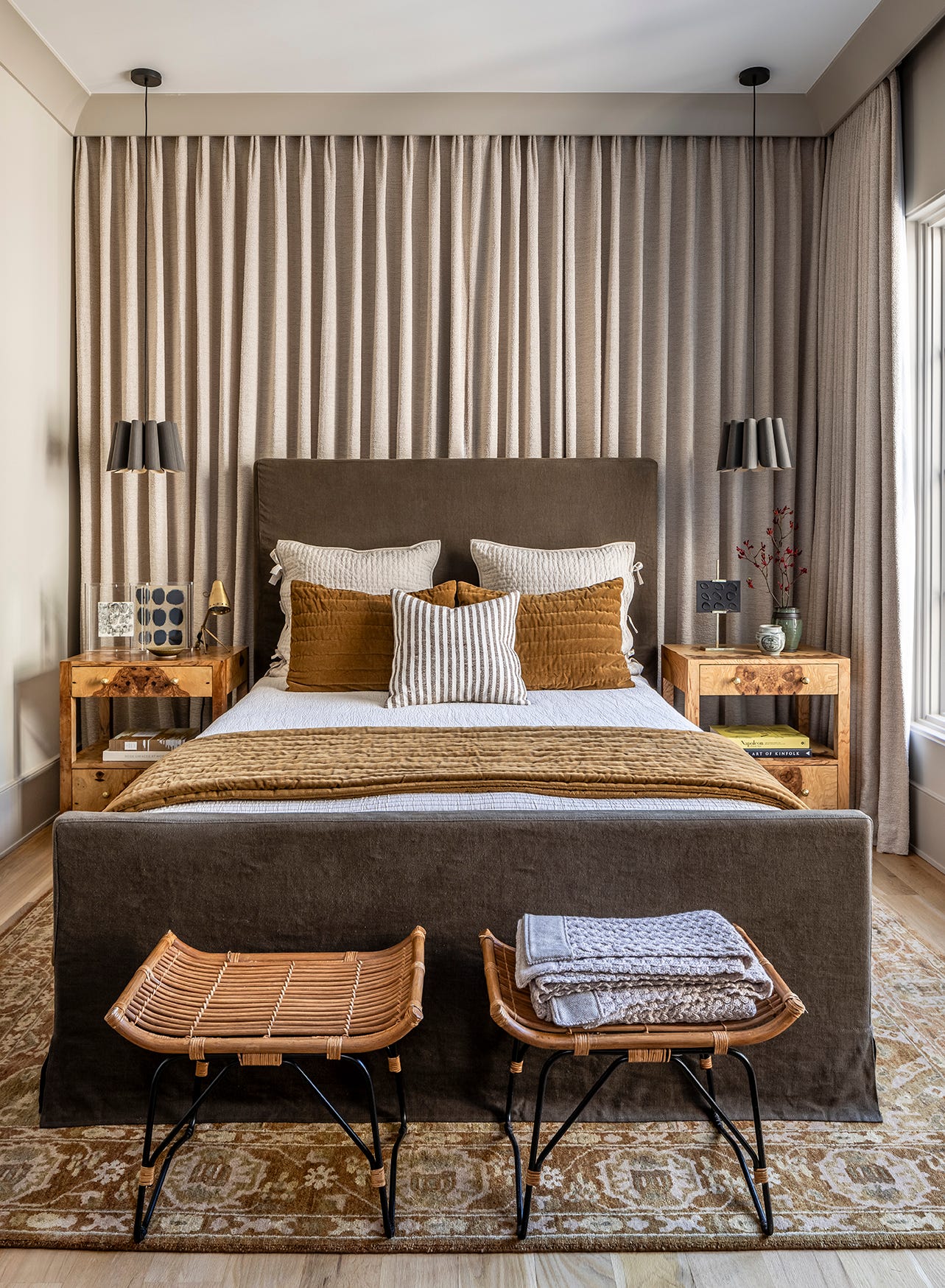 A symmetrical bedroom features a dark brown upholstered bed flanked by burl wood nightstands against a wall of floor-to-ceiling pleated curtains.