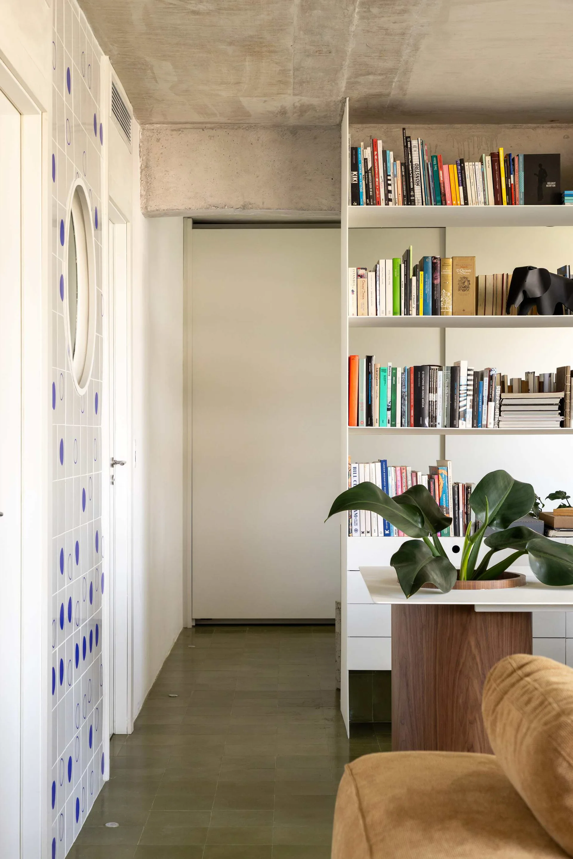A bright modern interior featuring an open white bookshelf filled with books, a blue patterned tiled wall, and an exposed concrete ceiling.