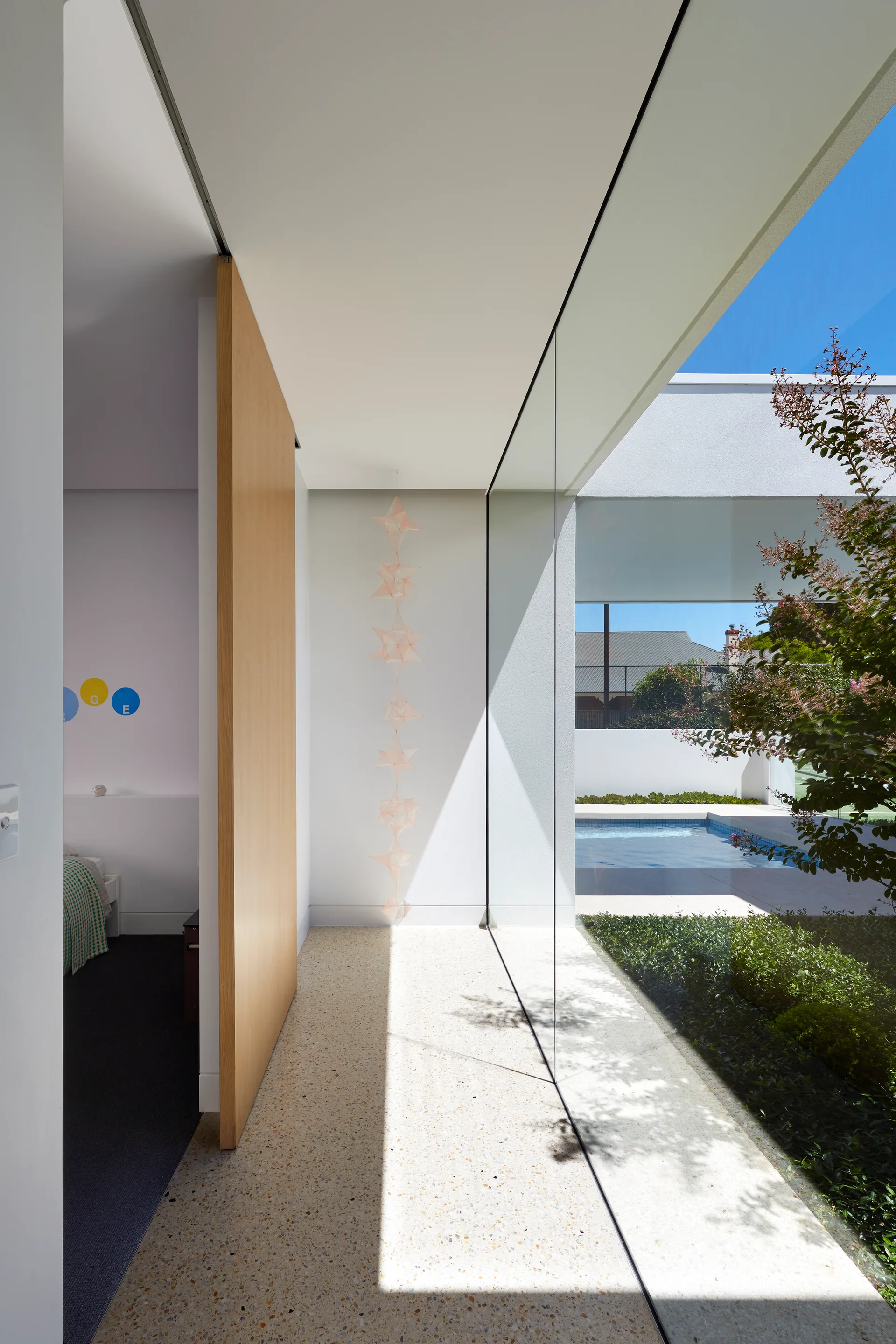 A minimalist hallway featuring terrazzo flooring, a large light-wood pivot door, and a floor-to-ceiling glass wall overlooking an outdoor pool.