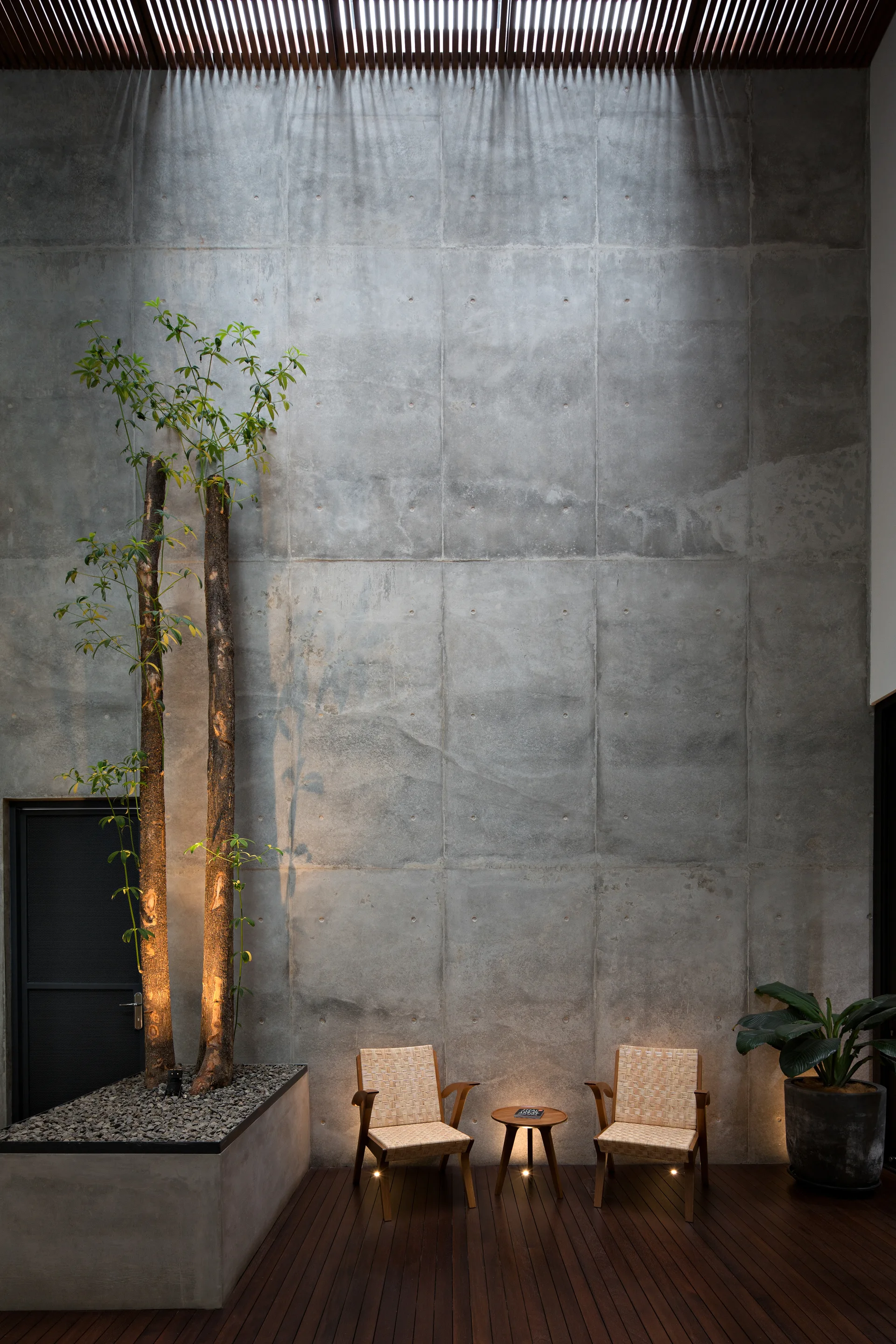 A modern outdoor patio features a tall concrete panel wall, dark wood decking, two woven armchairs with a small table, and built-in planters with trees.