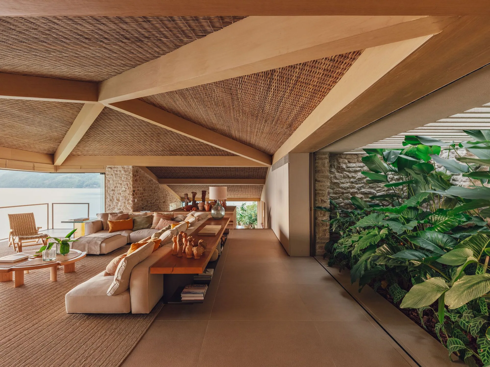 A spacious living room features a cream sectional sofa, a sloped wood-beamed ceiling with woven panels, and a garden bed of indoor tropical plants.
