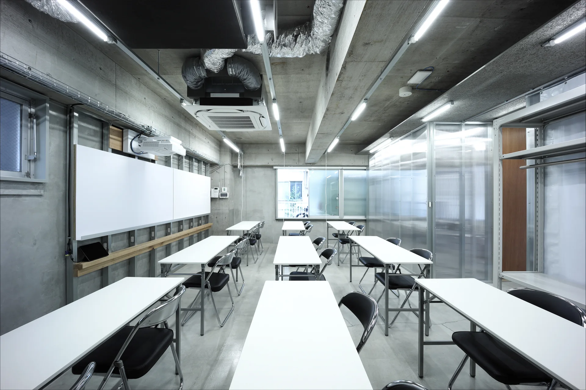Rows of white folding desks and black chairs face a whiteboard in an industrial room with raw concrete walls and an exposed ceiling.