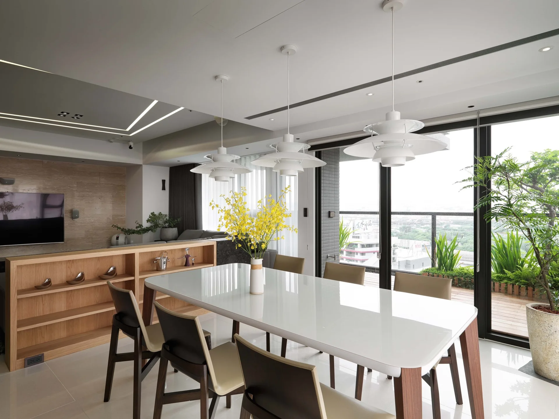 A contemporary dining room features a glossy white table, three layered pendant lights, and a low wooden bookcase next to sliding balcony doors.
