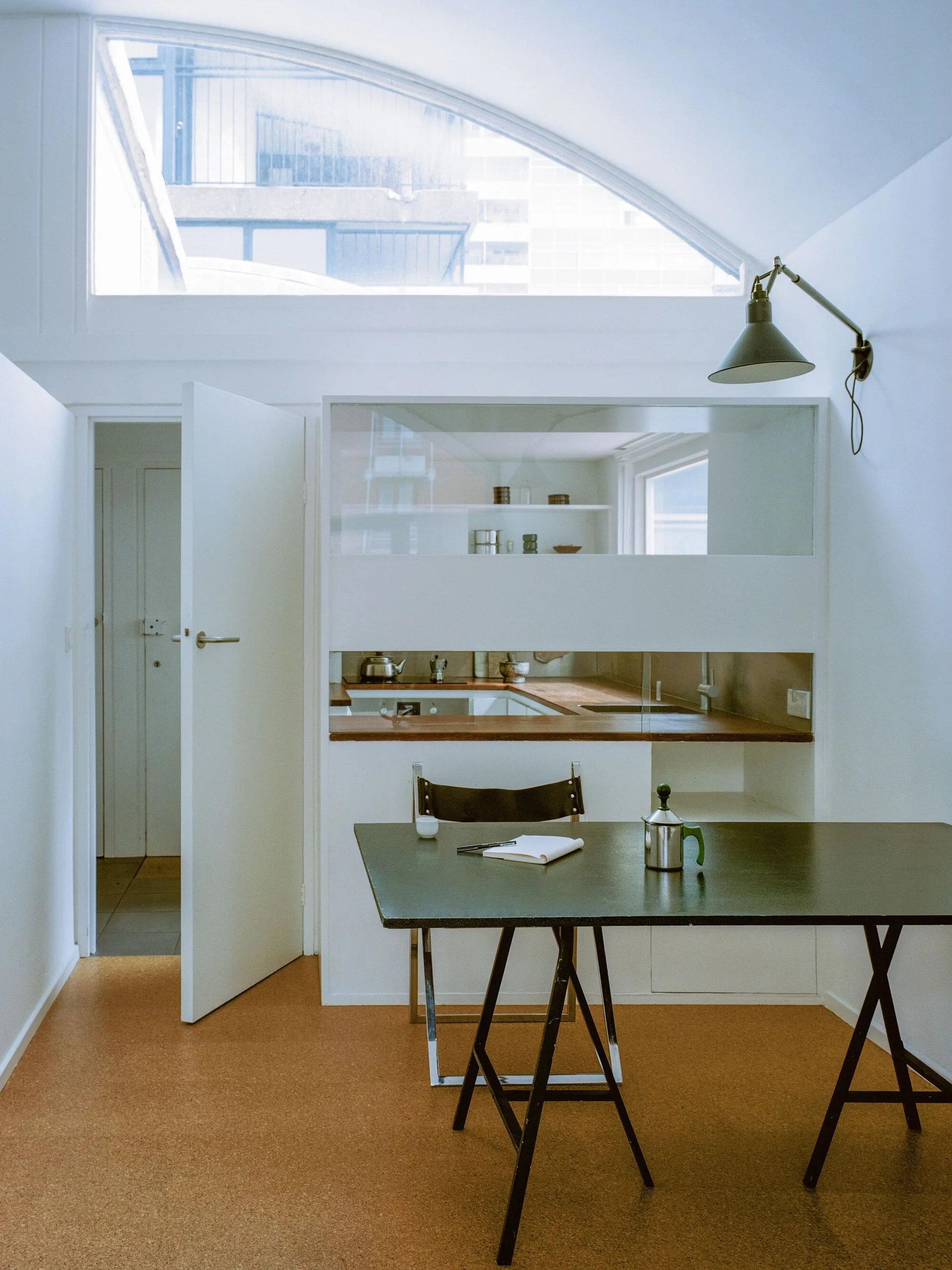 A minimalist dining table with a folding chair sits in front of a white built-in kitchen under a large arched clerestory window.
