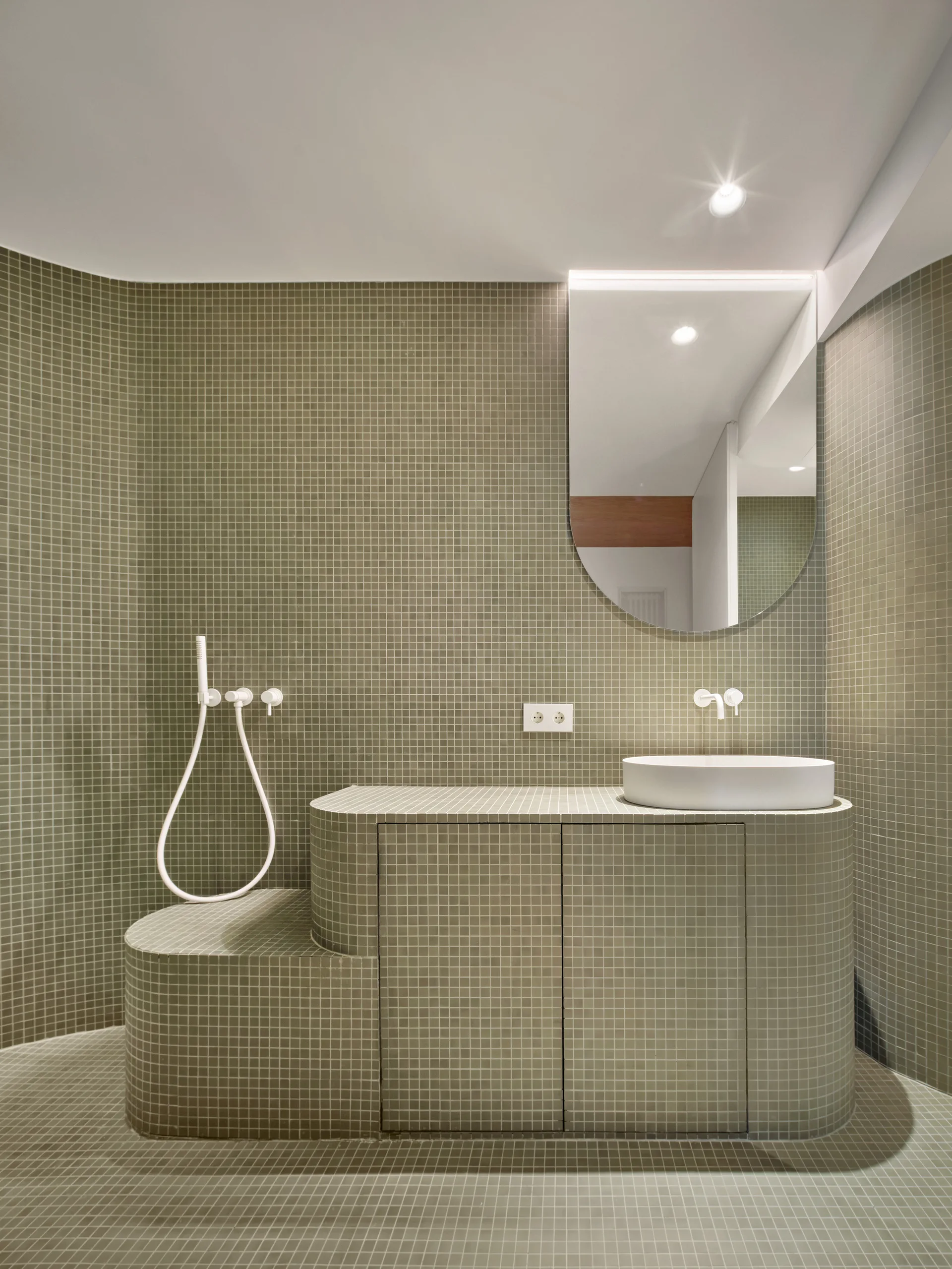A sage green bathroom covered in small grid mosaic tiles features a curved integrated vanity, white vessel sink, and an arched mirror.