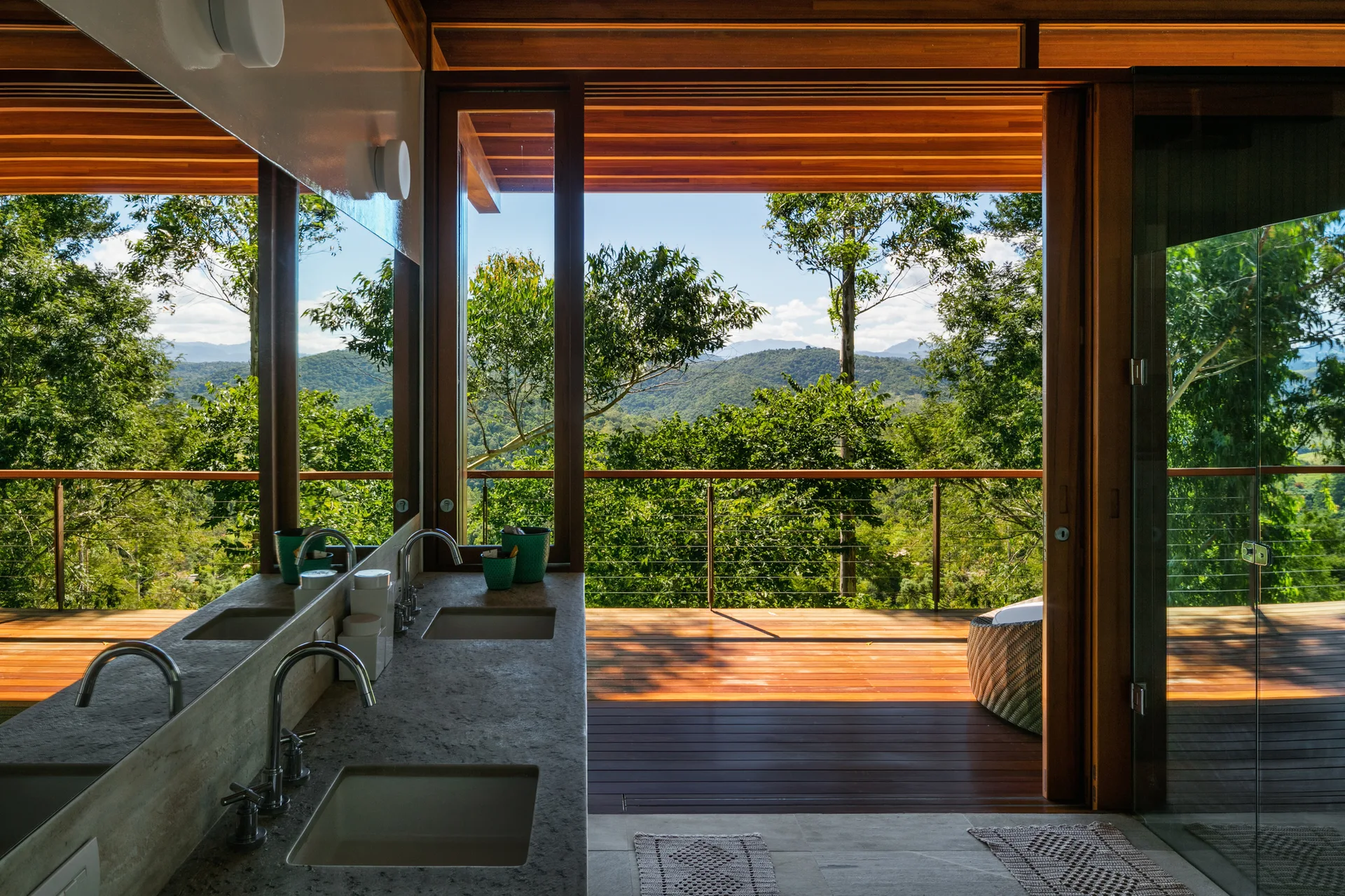 A stone double vanity with chrome faucets sits inside a bathroom that opens onto a wooden deck overlooking a lush green forest.