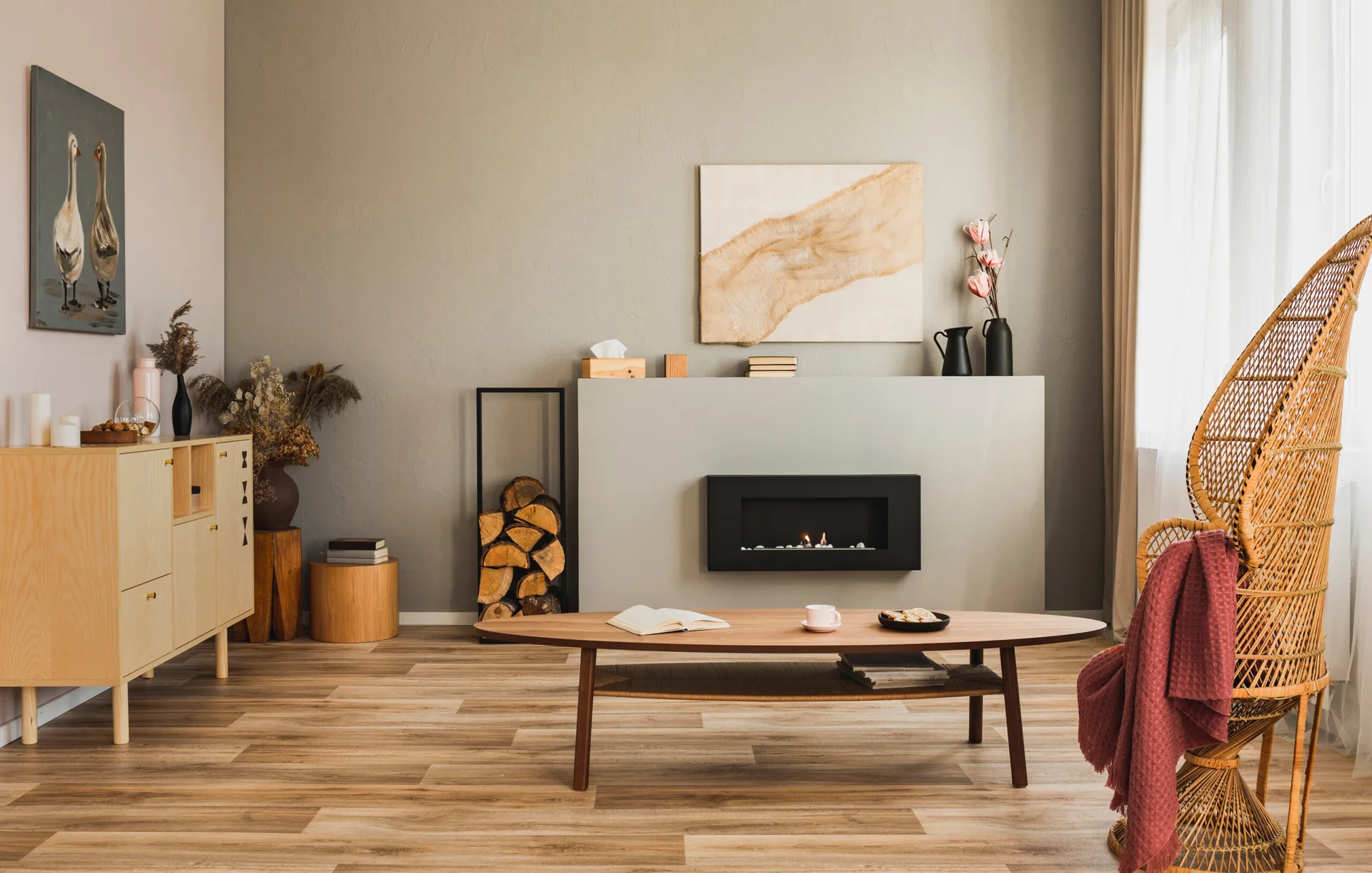 A minimalist living room featuring a black inset fireplace, a wooden oval coffee table, and a tall woven rattan armchair beside a window.