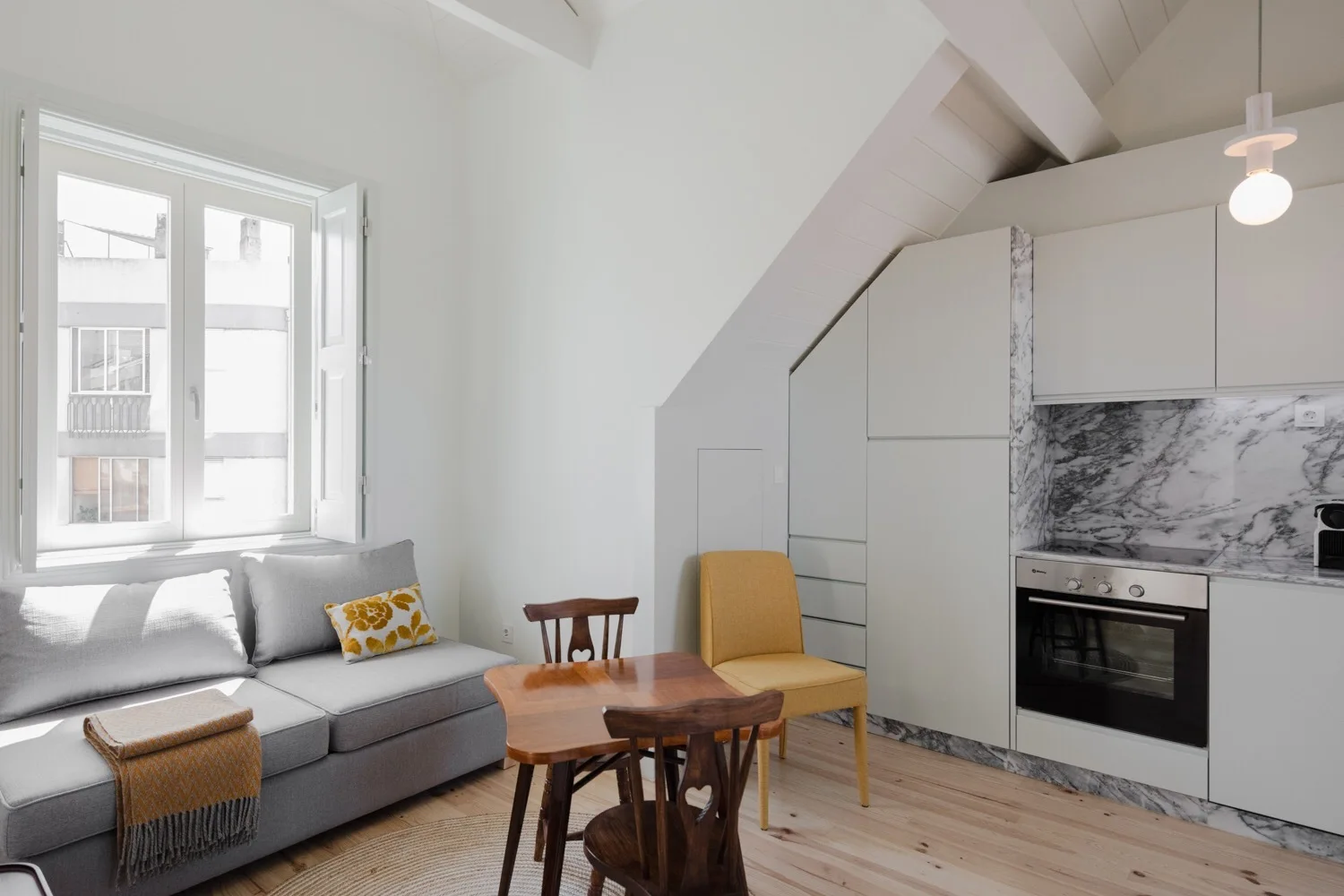 A compact attic kitchen and living space features custom white cabinets under a sloped ceiling, a grey sofa, and a marble backsplash.