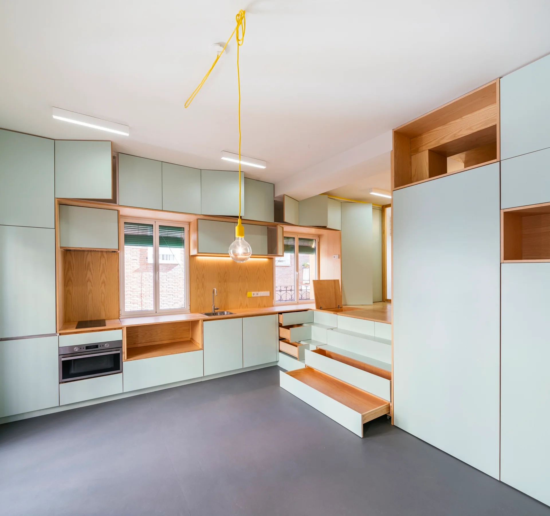 A minimalist kitchen features pale green modular cabinets, light wood trim, and integrated stepped storage drawers leading to an upper level.