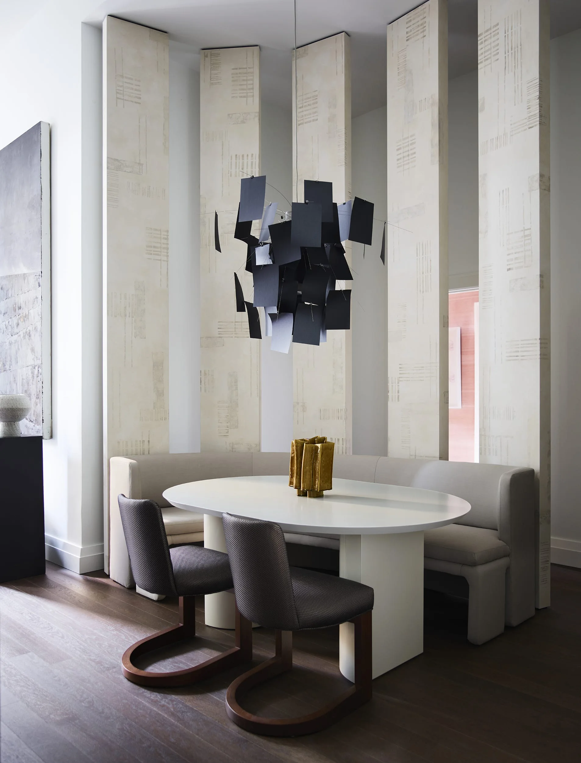 A modern dining area features a curved beige banquette, an oval white pedestal table, and a black sculptural pendant light against patterned wall panels.