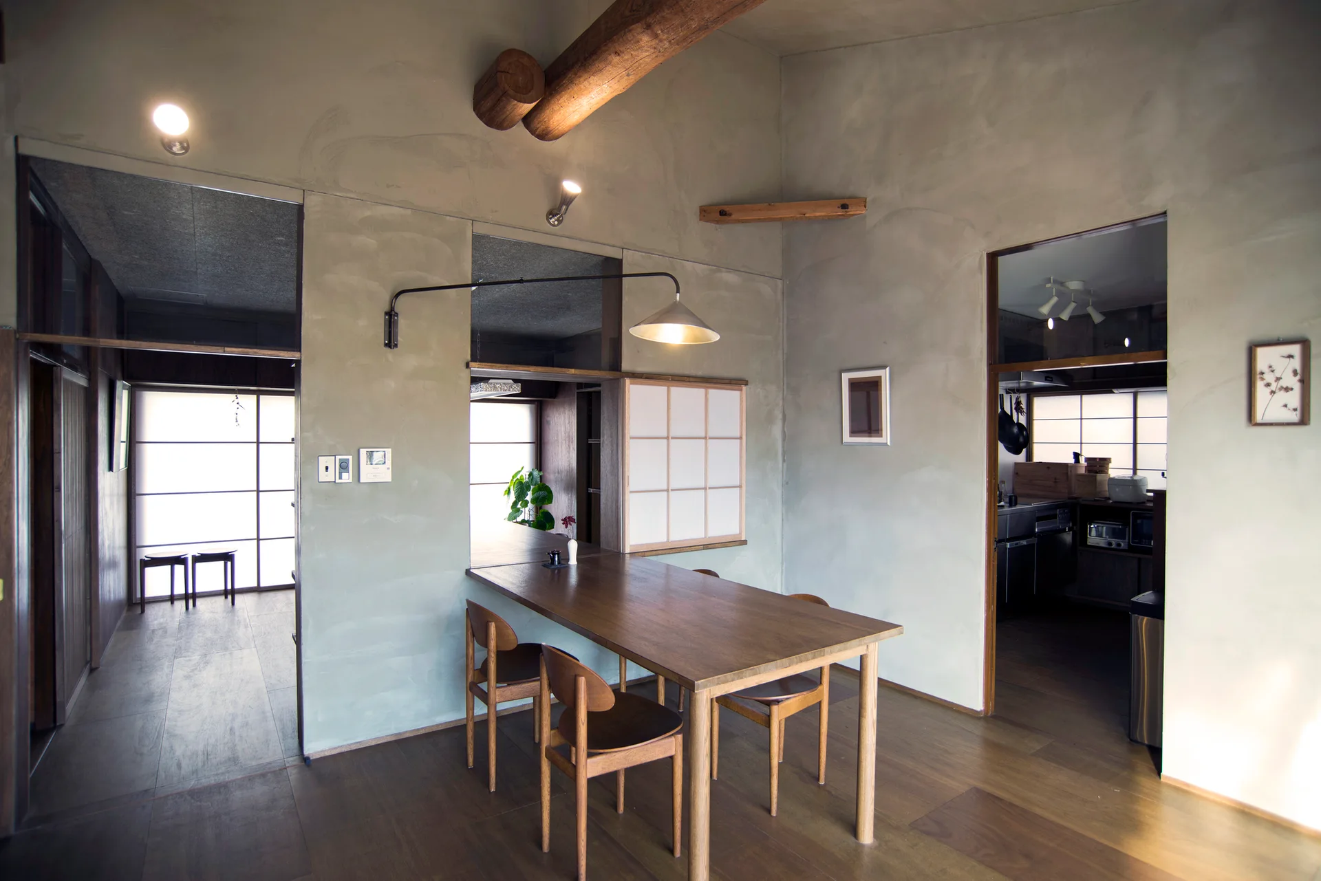 A minimalist dining room features a dark wood table and matching chairs set against grey plaster walls with an exposed timber ceiling beam.