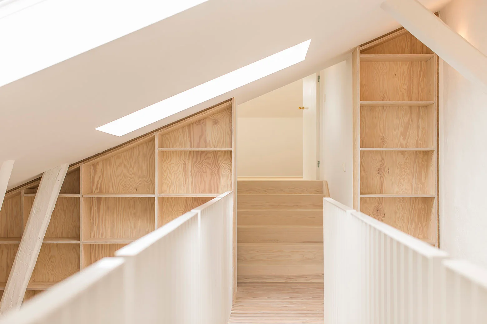 A bright attic landing features custom plywood bookshelves built into the sloped walls, white slatted railings, and wooden stairs.