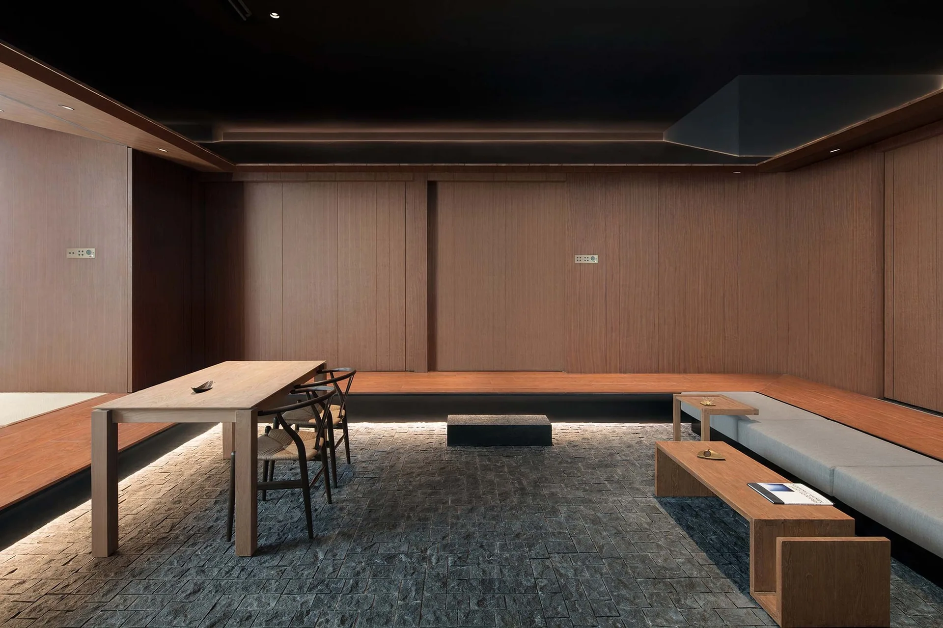 A minimalist room featuring a dark stone paver floor, a raised wooden platform with under-cabinet lighting, a dining table, and a grey bench.