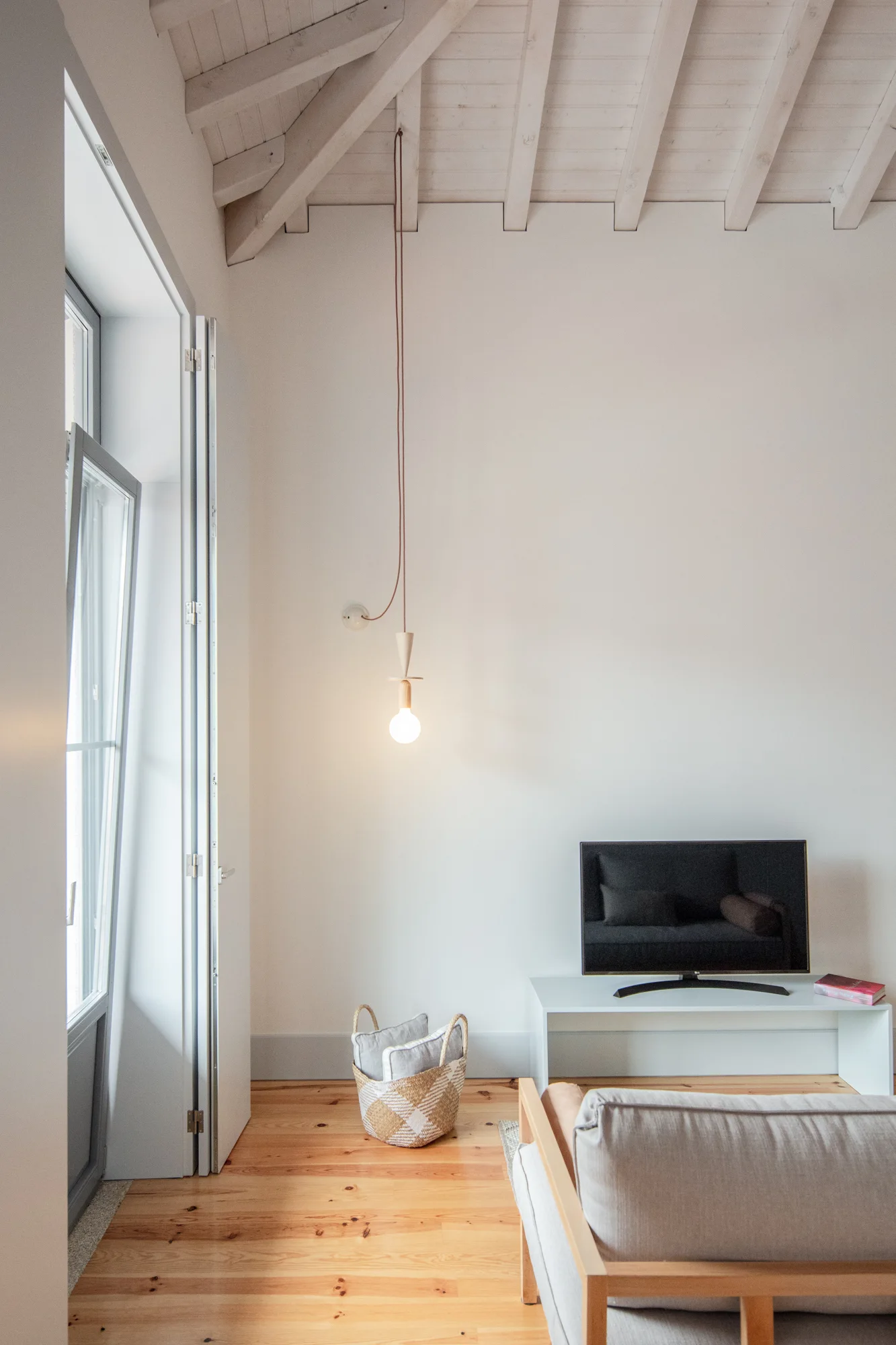 A minimalist living room with a low white console, hanging pendant light, light wood floor, and white painted ceiling beams.