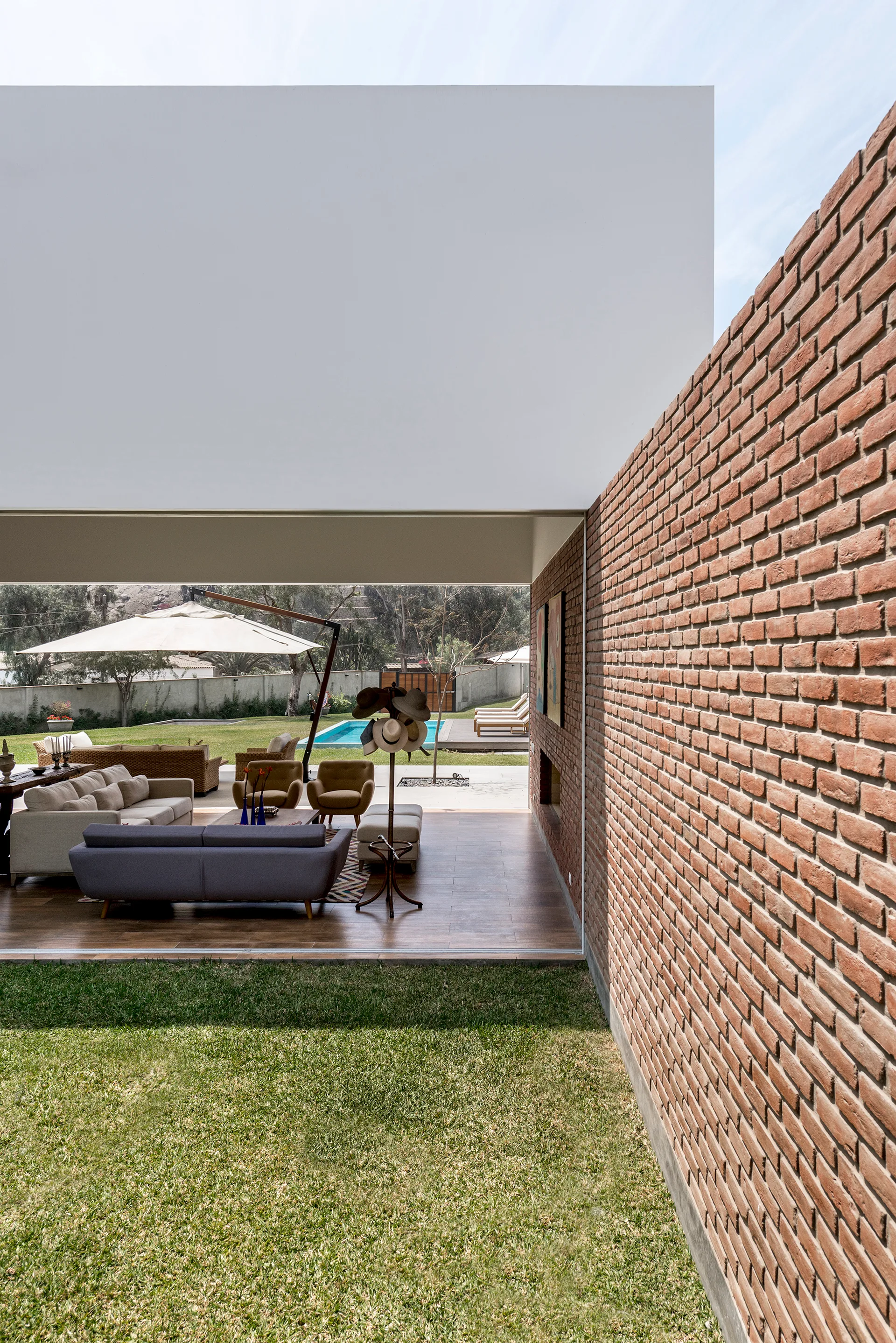 An open-sided modern living pavilion with neutral sofas next to a long red brick wall leading out to a grassy lawn and swimming pool.