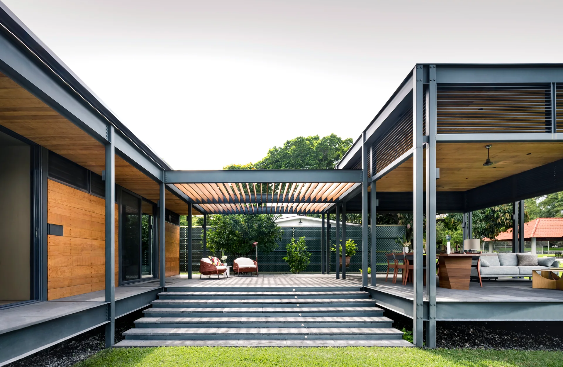 An elevated modern outdoor patio features a dark steel structural frame, warm wood ceilings, wide grey stone steps, and outdoor lounge furniture.
