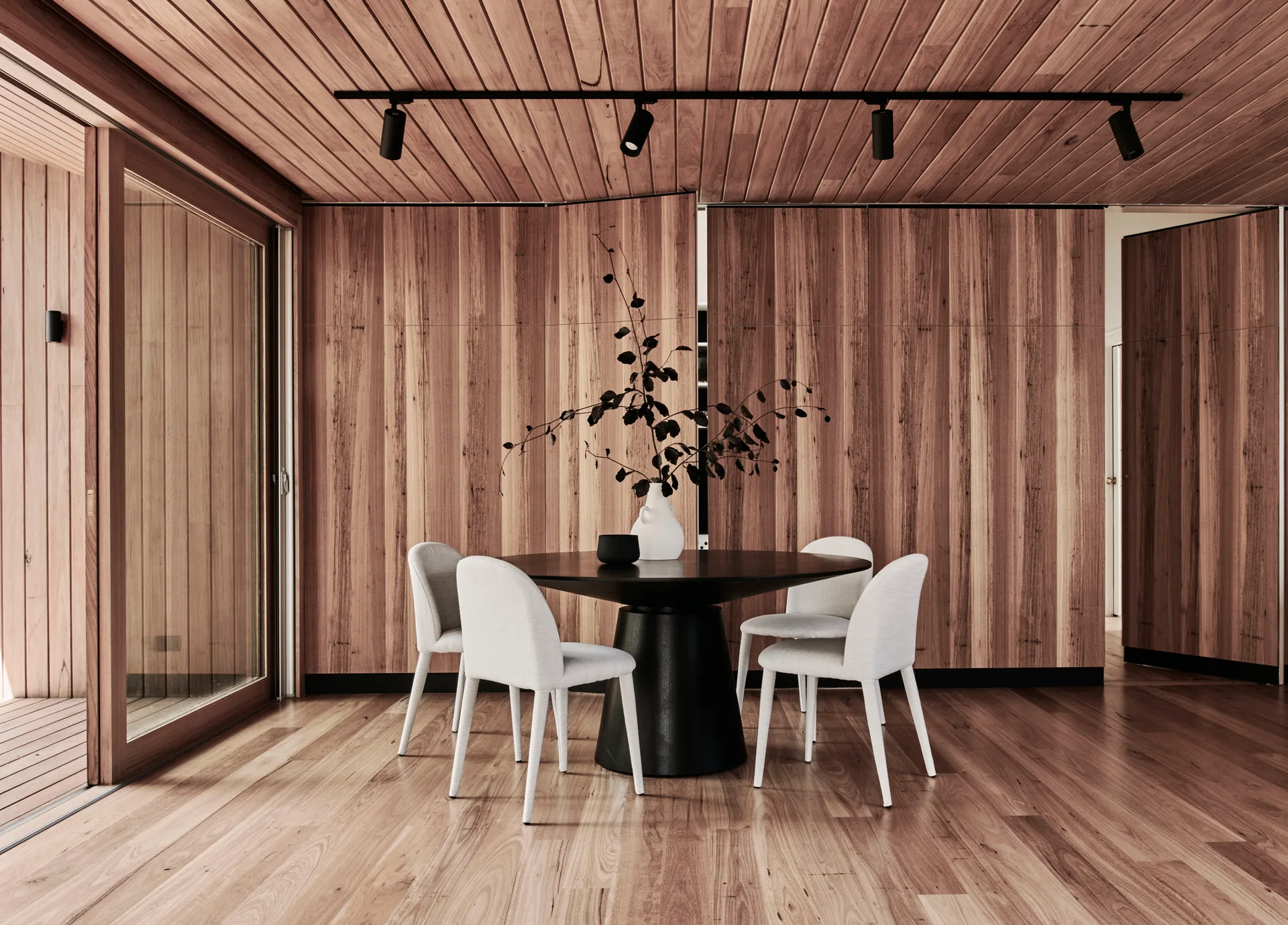 A round black dining table and four light gray chairs sit in a room clad entirely in vertical wood paneling, under a planked ceiling with track lights.