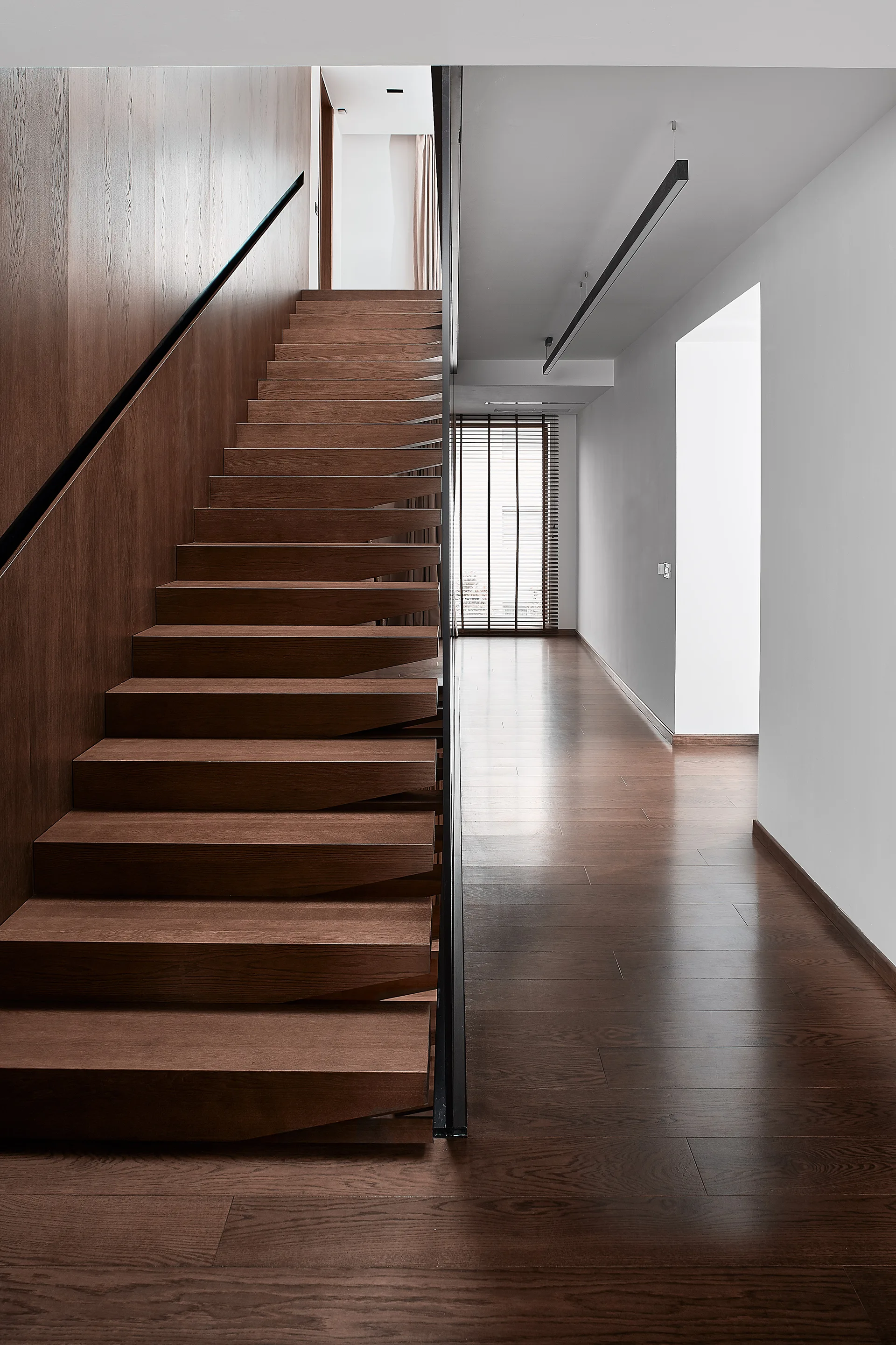 A modern wooden staircase with matching wood wall paneling next to a hallway with white walls and a linear ceiling light fixture.
