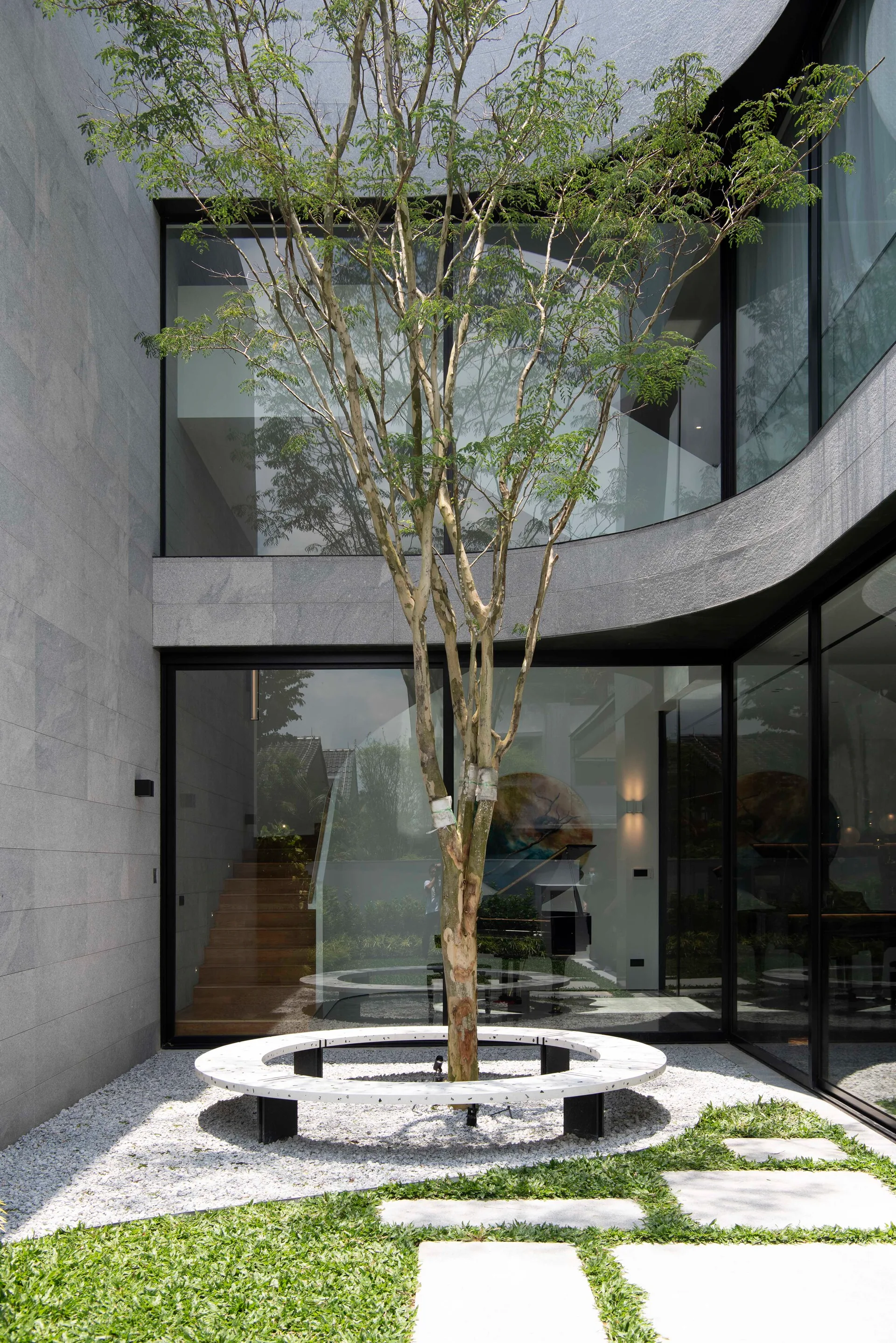 A modern residential courtyard features a central tree encircled by a white bench, set against large glass windows and grey stone walls.