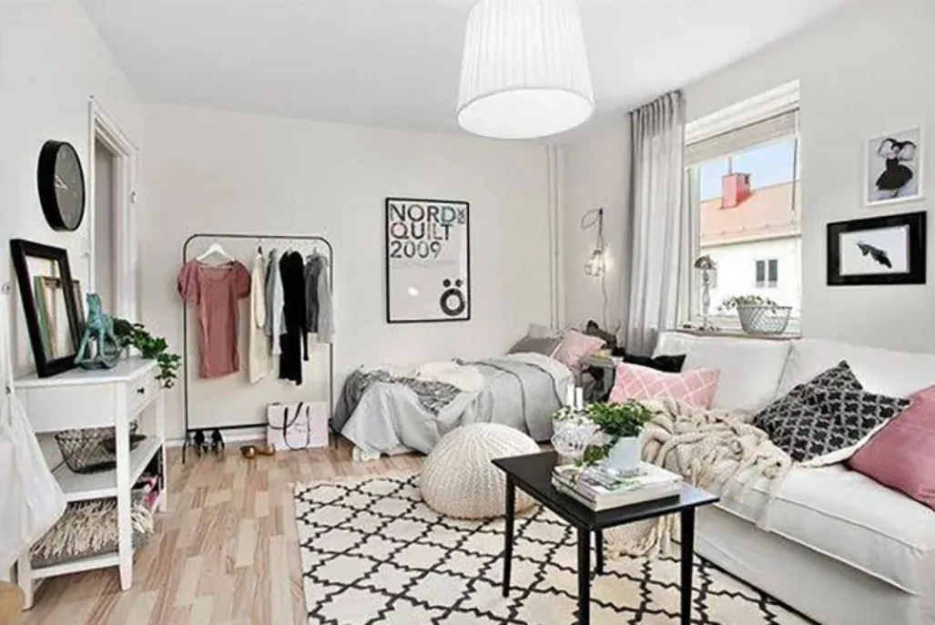 A Scandinavian studio apartment features a combined bedroom and living area with a white sofa, a bed, a black coffee table, and a clothes rack.
