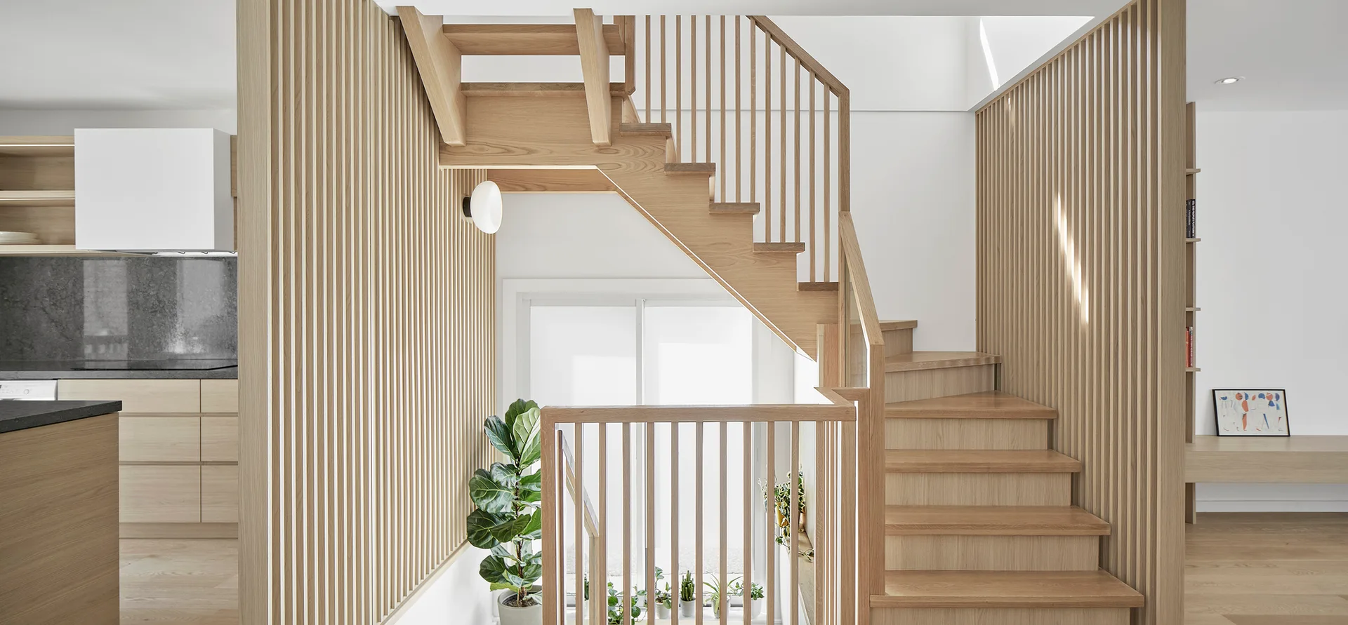 A light wood open-riser staircase with vertical slat paneling stands next to a kitchen with matching cabinetry and a large window.
